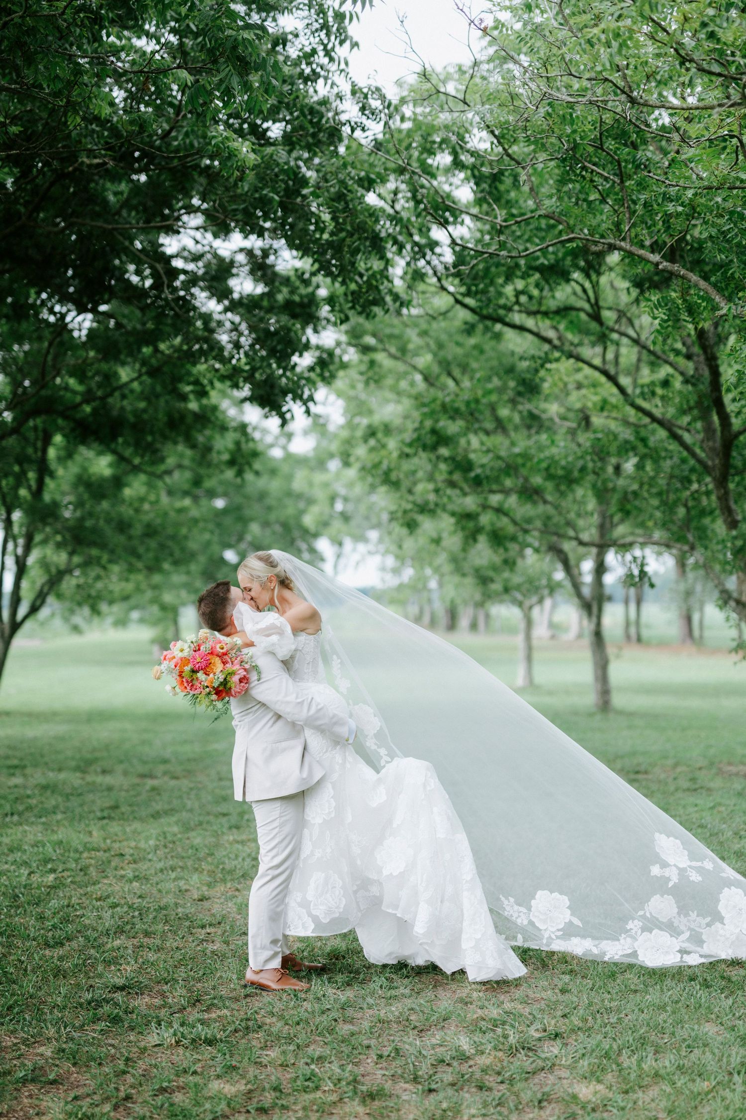 Bride and groom share a romantic kiss outdoors, with her lace veil flowing in the breeze amid lush green trees.