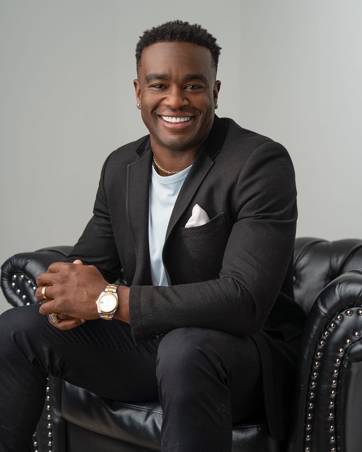 Smiling man in black blazer and white shirt sitting on leather chair, wearing gold watch and ring.