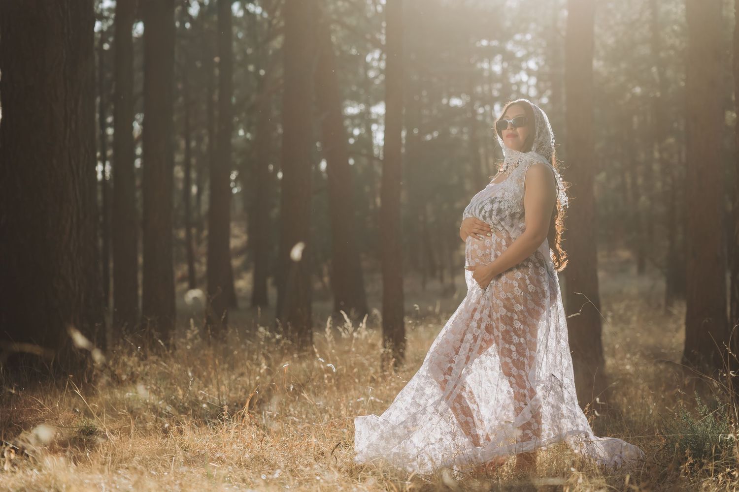 Pregnant woman in lace gown posing in sunlit forest for maternity photography session.