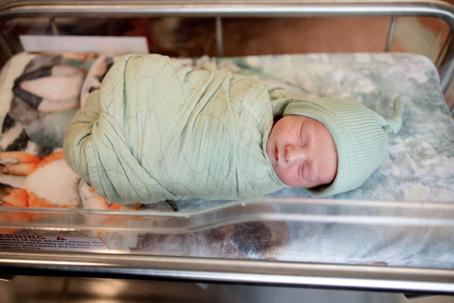Newborn baby swaddled in green blanket and hat sleeping peacefully in hospital bassinet.