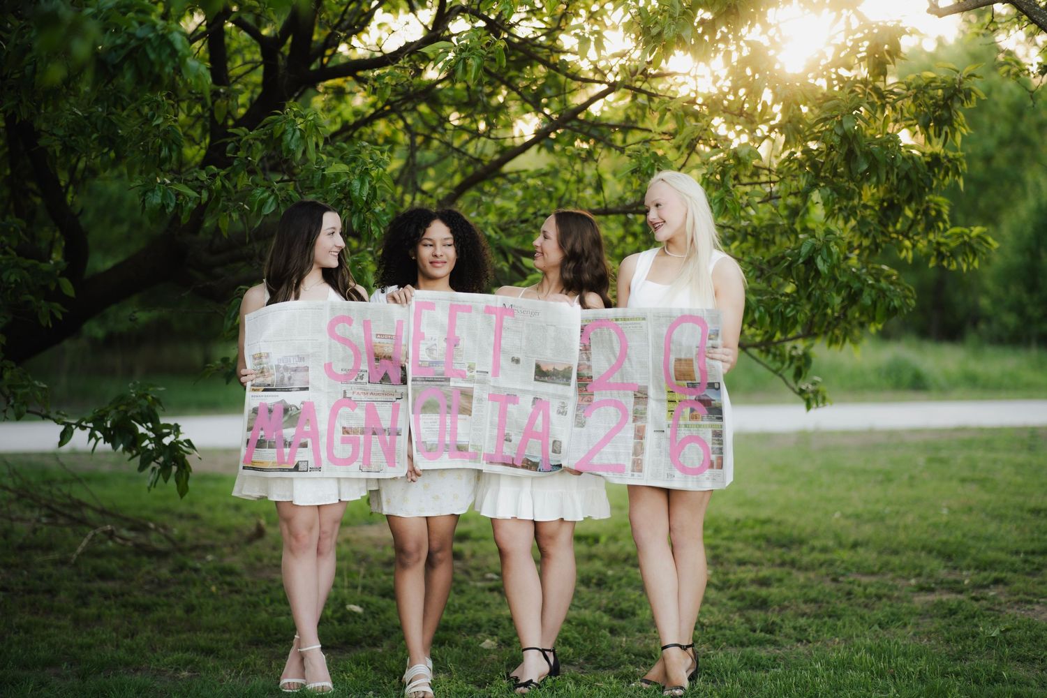 Four young women holding a pink decorative banner together in a sunlit park.