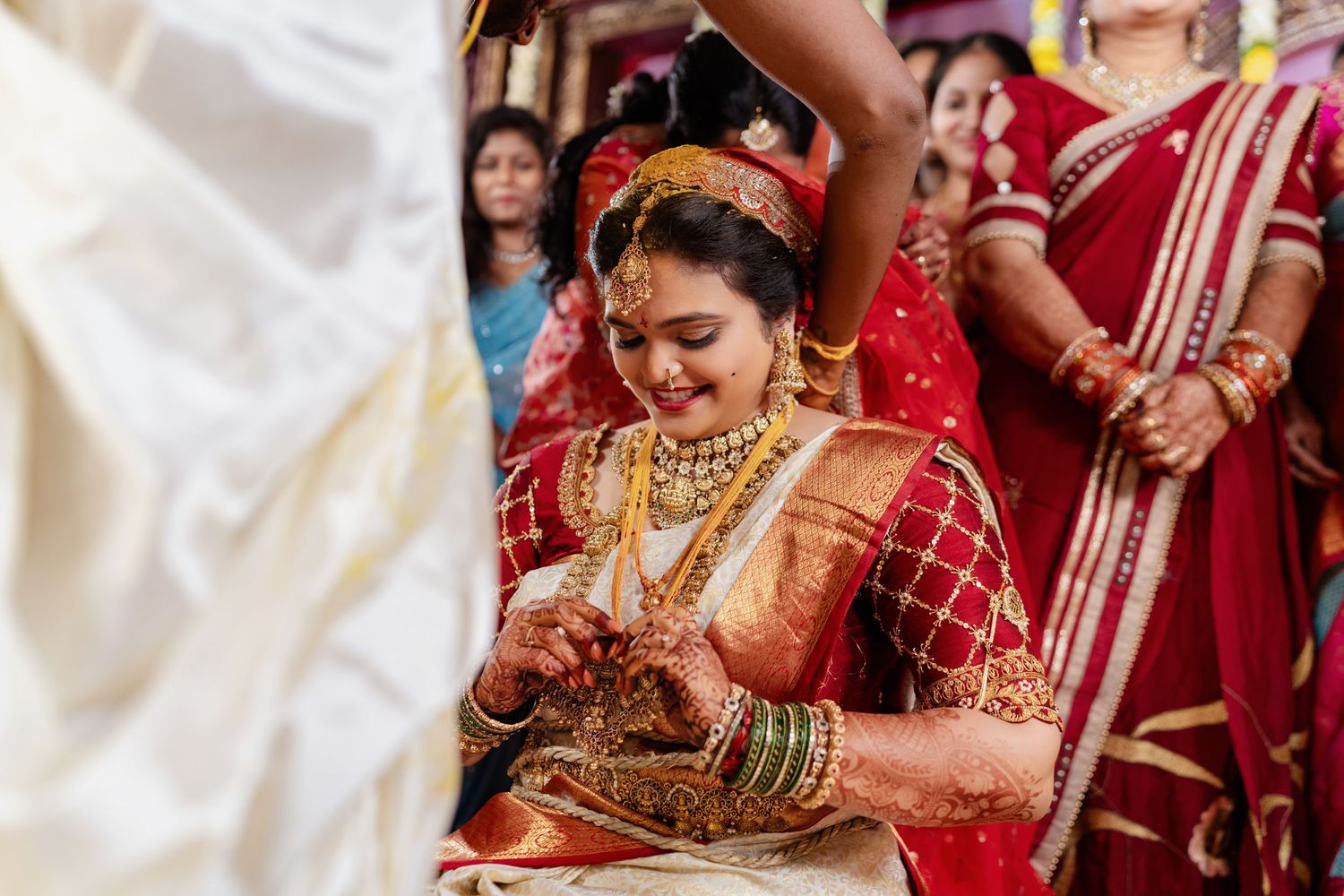 A bride in a red and gold saree adorned with traditional jewelry smiles during a South Indian wedding ceremony.