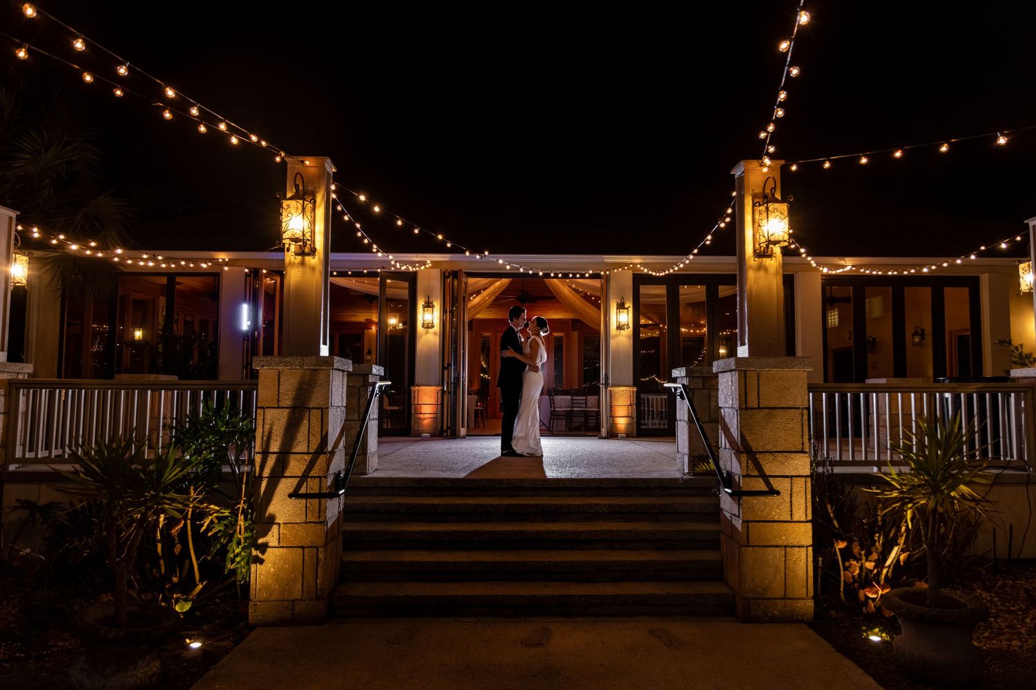 Couple sharing a romantic kiss on illuminated venue steps at night, surrounded by twinkling string lights.