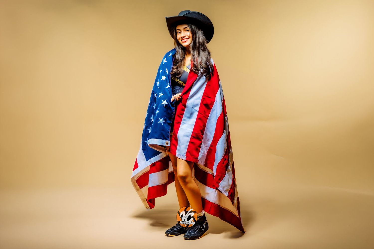 Young woman in black hat wrapped in American flag, posing confidently against a beige background.