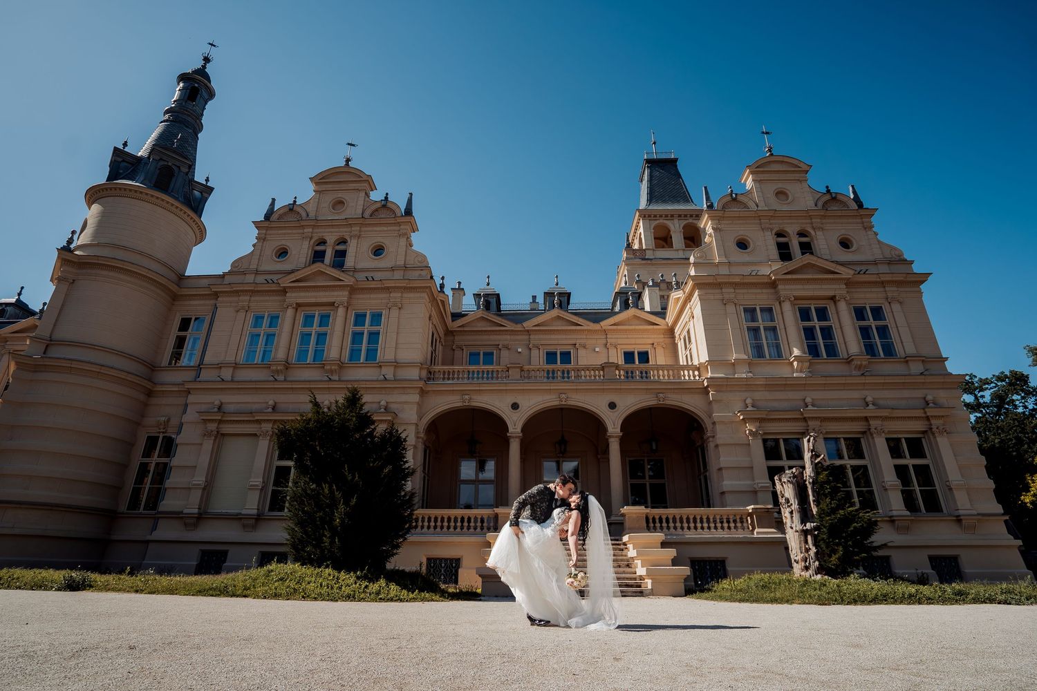 A menyasszony és a vőlegény romantikus csókot váltanak egy elegáns barokk palota előtt egy napsütéses esküvőn.