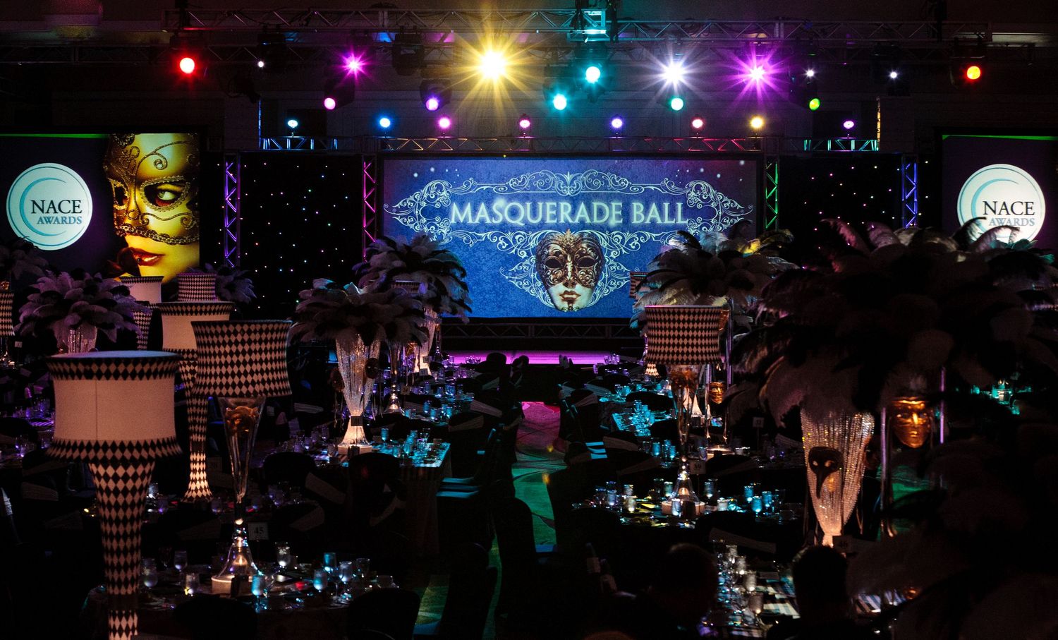 Elegant masquerade ball event with feathered centerpieces, dramatic lighting, and a glowing stage backdrop in a dark ballroom.