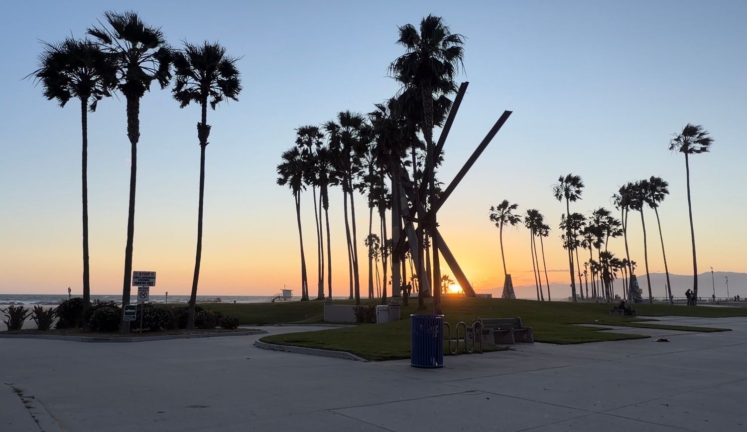 Sunset silhouette of tall palm trees and a sculpture at Venice Beach, California, with golden sky reflecting on the horizon.