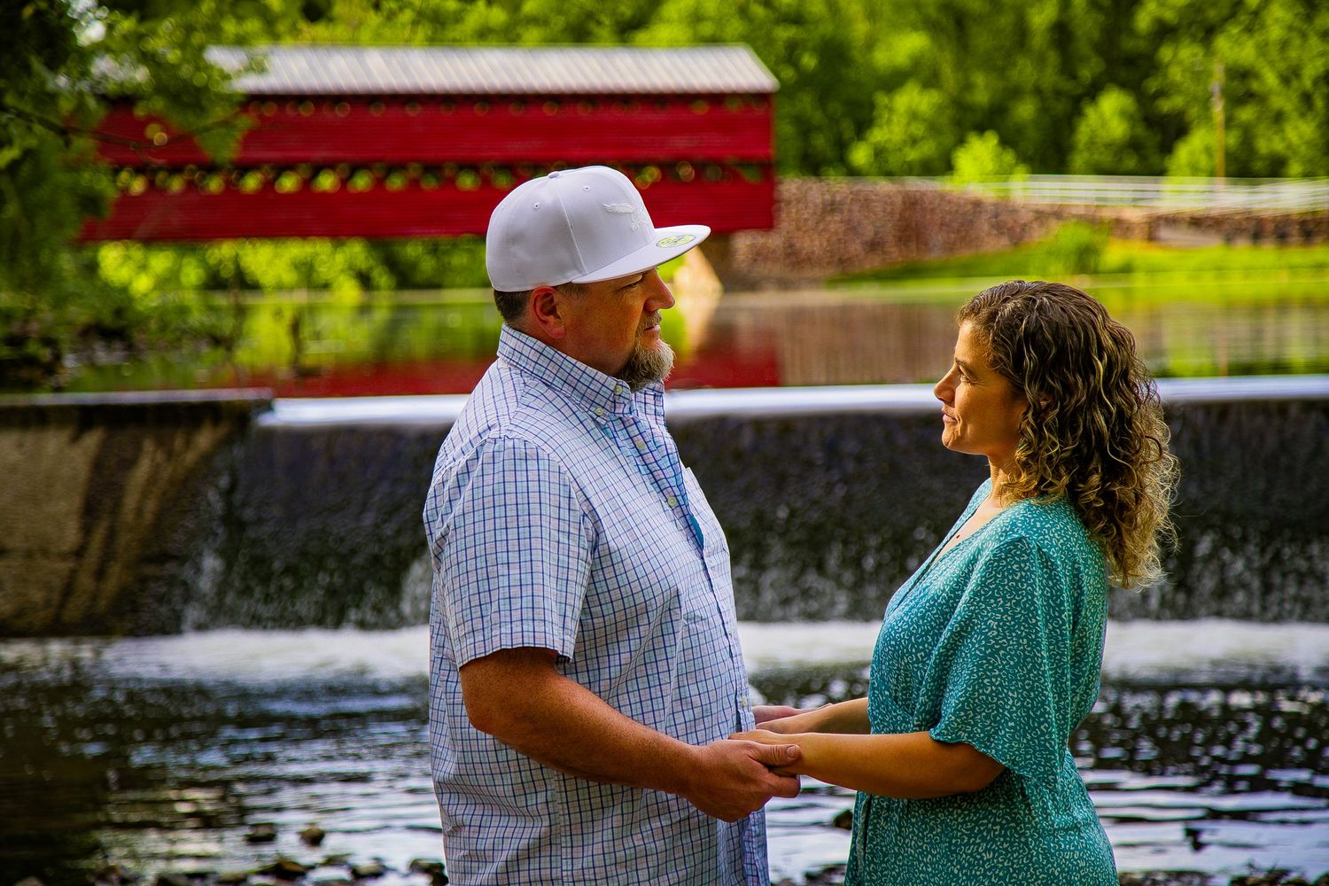 Couple holding hands by a scenic red covered bridge and waterfall surrounded by lush green trees.