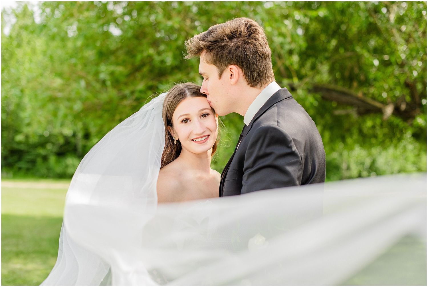 Bride and groom share a romantic moment outdoors, with her veil flowing in the breeze against lush green trees.