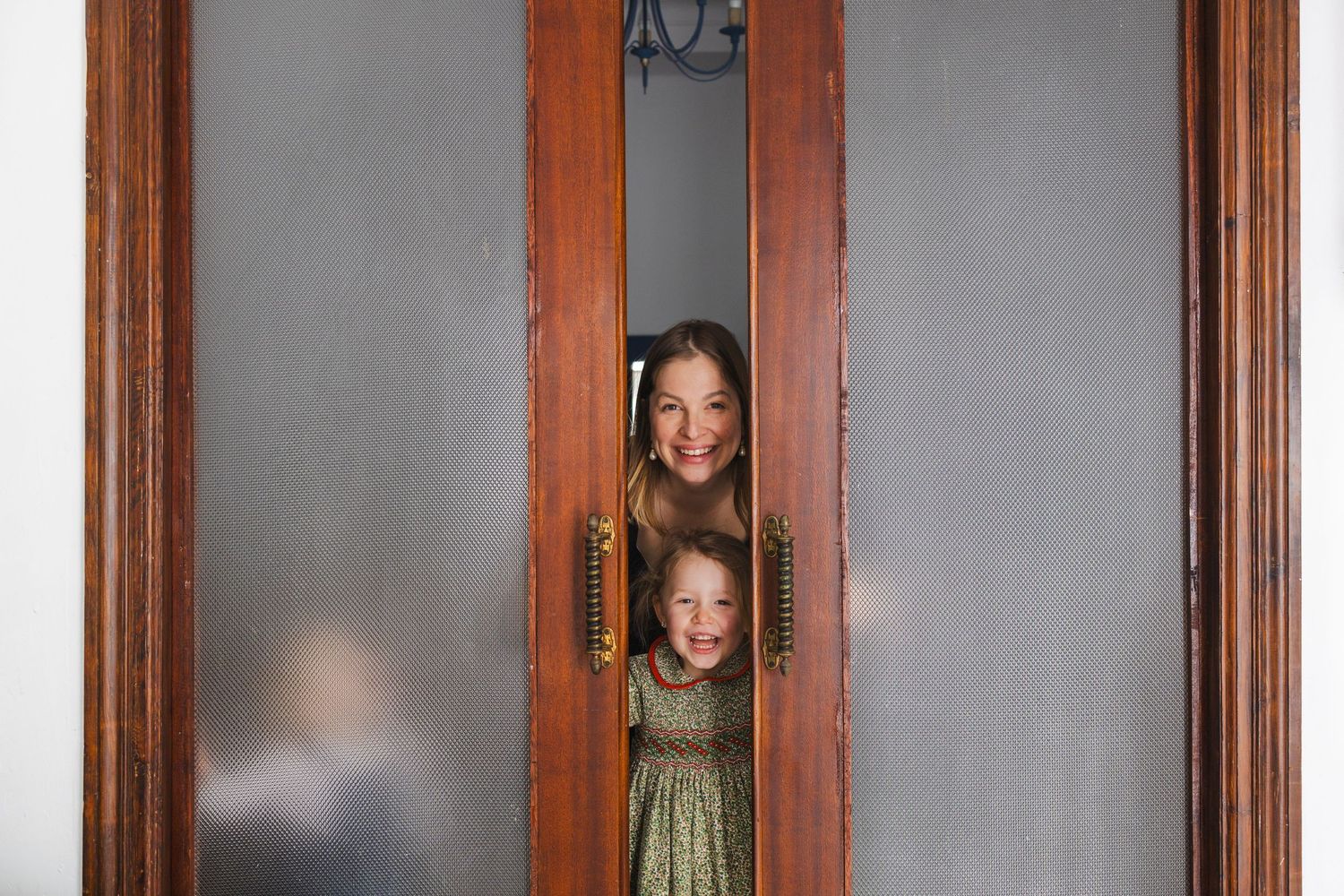 Child playing with billowing white curtains in a classic living room with checkered floors and antique wooden cabinet.