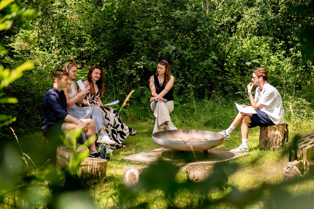 Small group of people sitting on logs around fire pit in lush woodland setting.