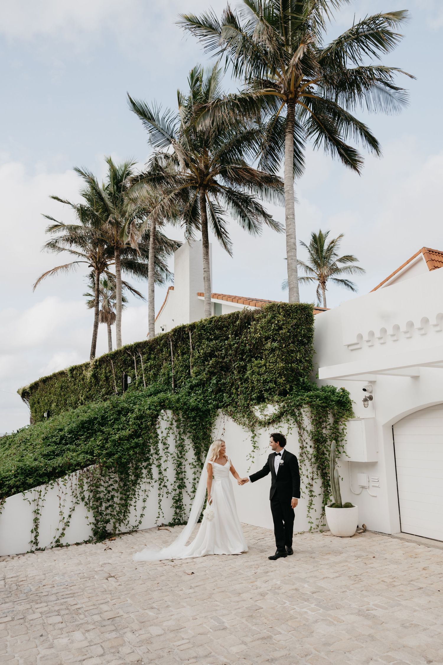 Bride and groom hold hands outside ivy-covered white estate with tall palm trees on their wedding day.