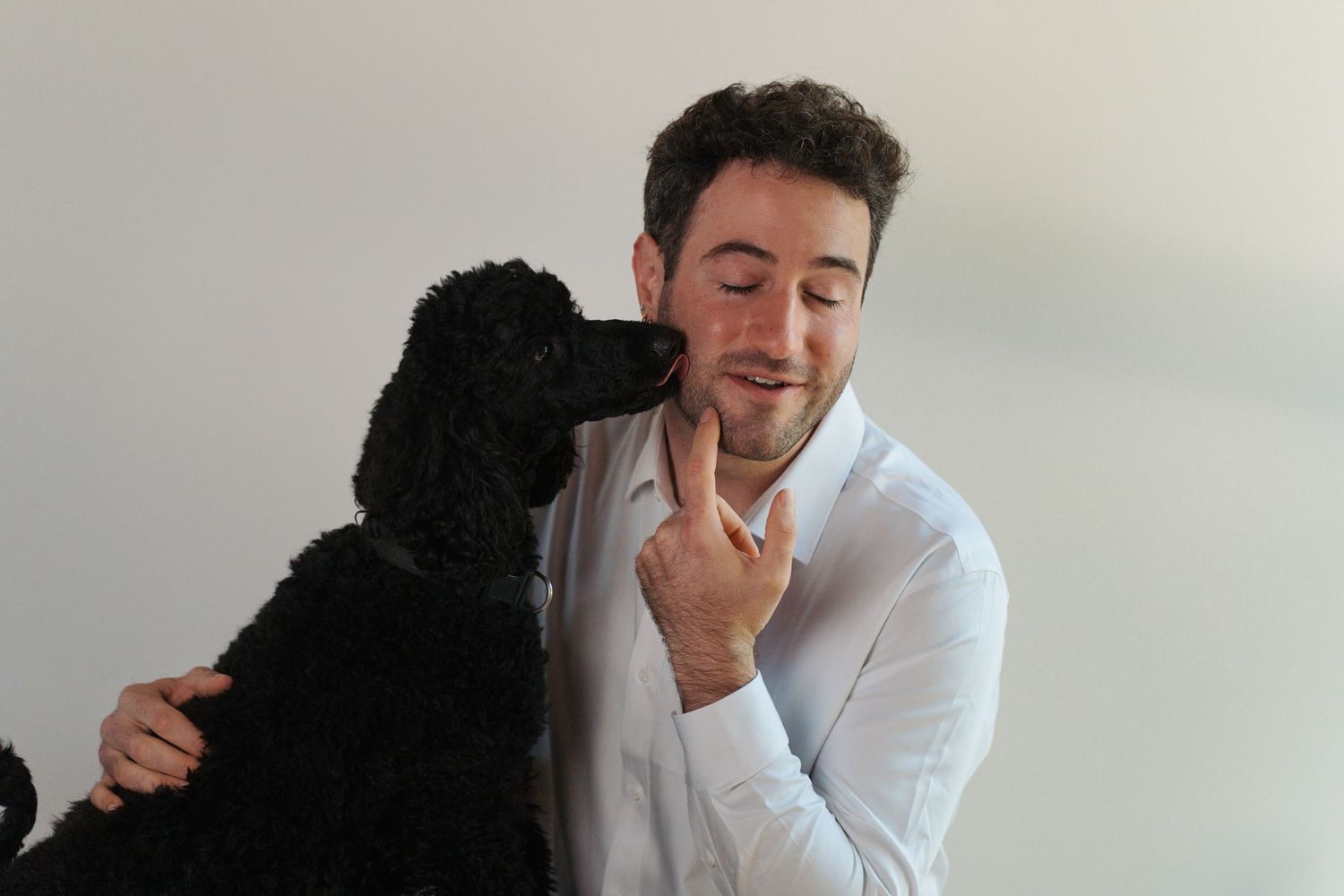 A young man in a white shirt smiles as a black poodle affectionately licks his face indoors photoshoot at Gables Hills district