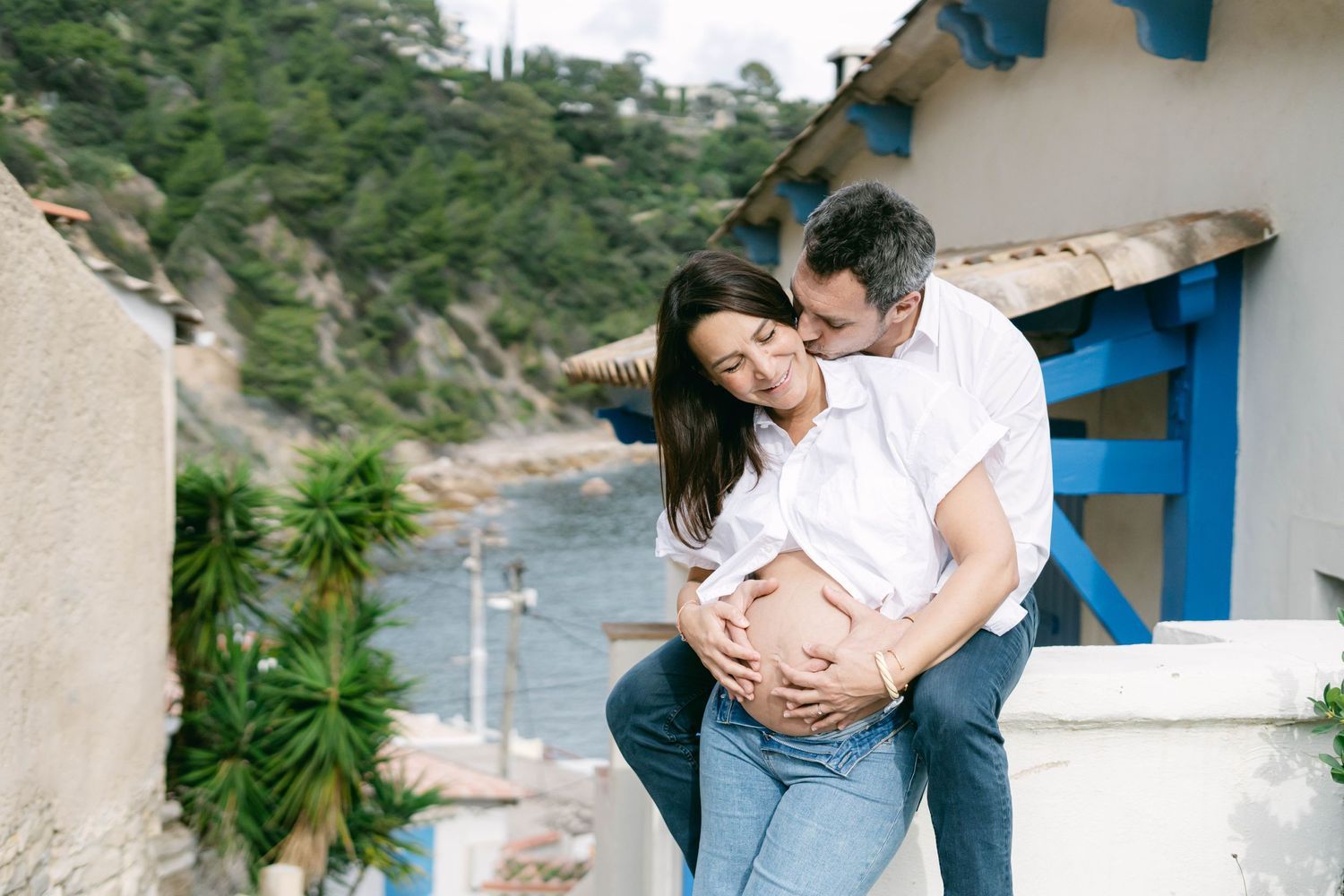 A couple embraces lovingly on a scenic outdoor terrace with lush greenery and mountains in the background.