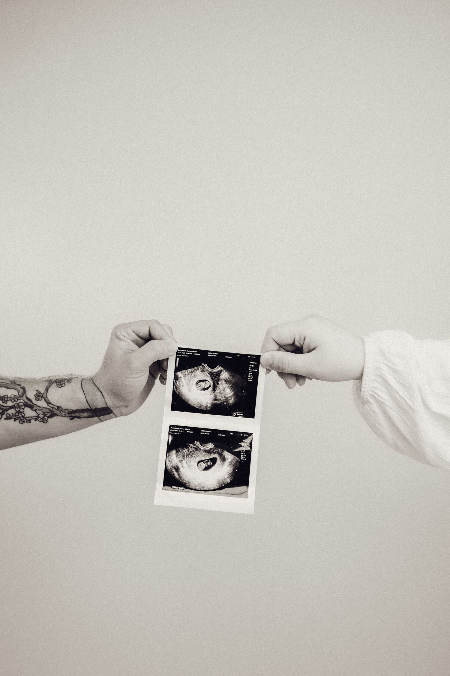 Two hands holding ultrasound photos, one with a tattoo, black and white image.