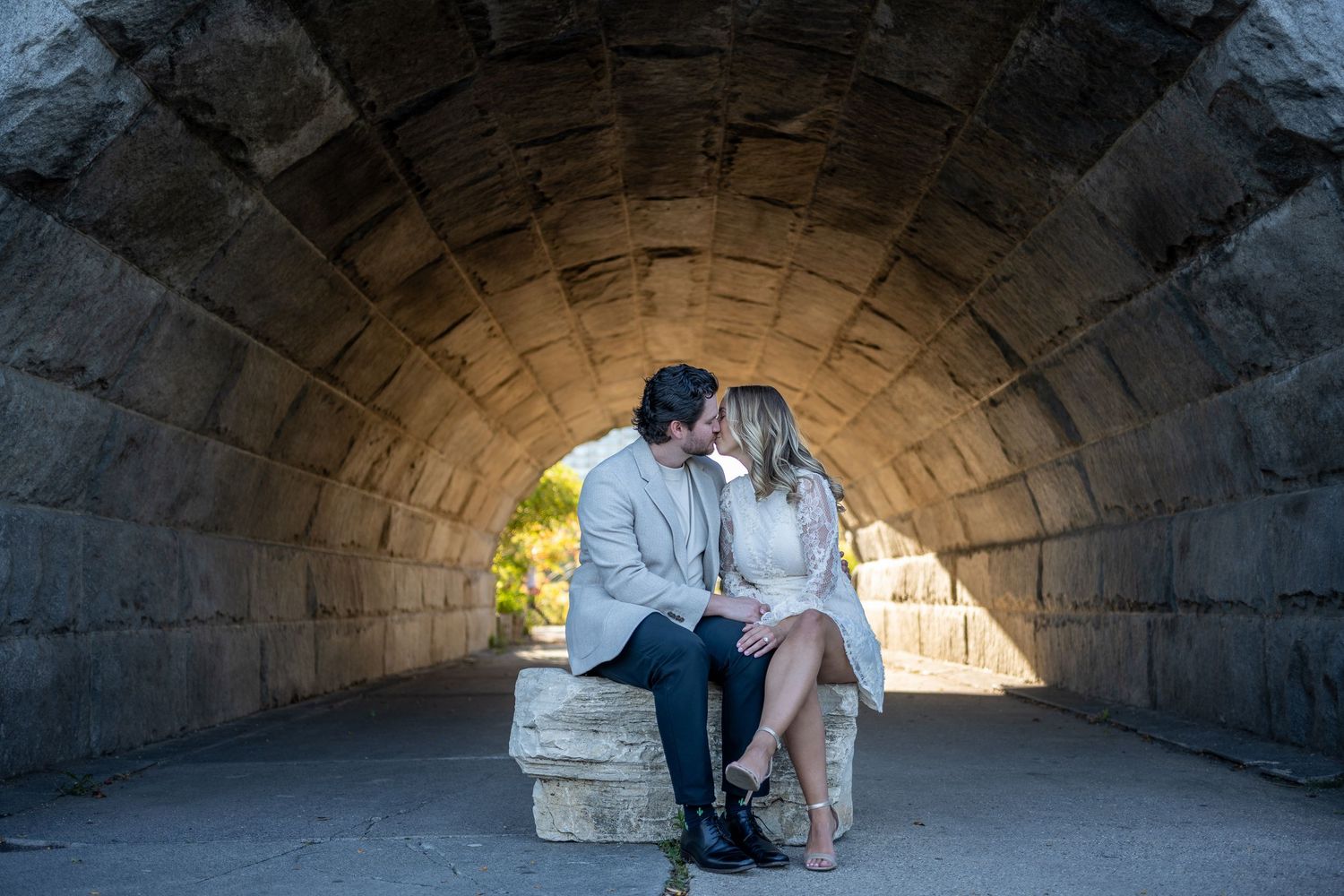 Couple sharing a romantic kiss under a stone archway tunnel, seated on a bench in Central Park.