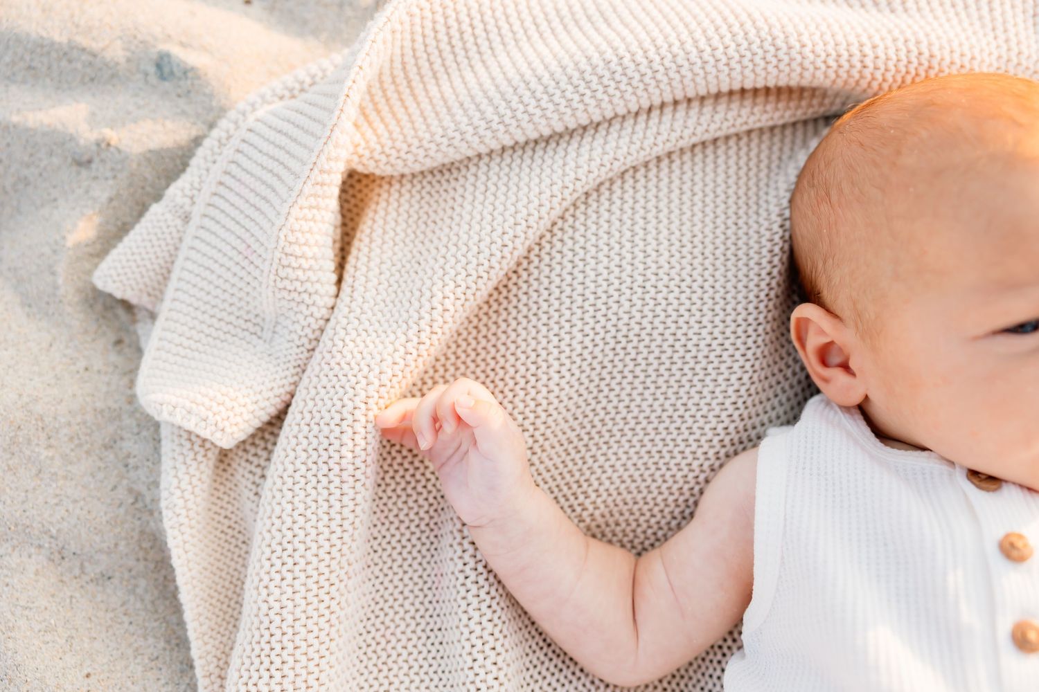 A peaceful baby resting on a soft beige knit blanket on sandy beach, wearing a white onesie.