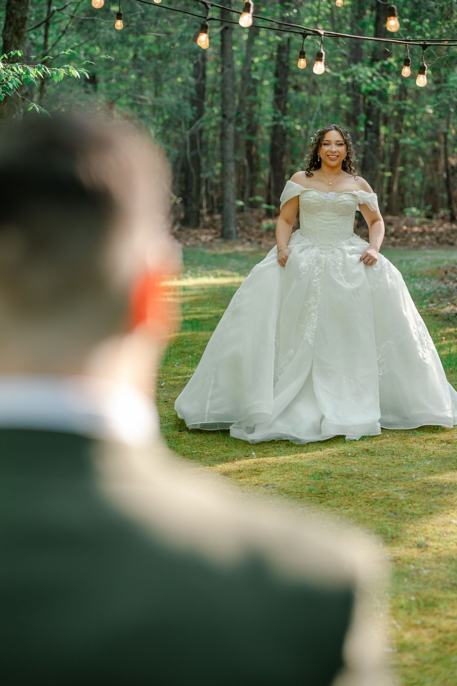 Bride in elegant off-shoulder ball gown smiles at groom during outdoor forest wedding first look ceremony.