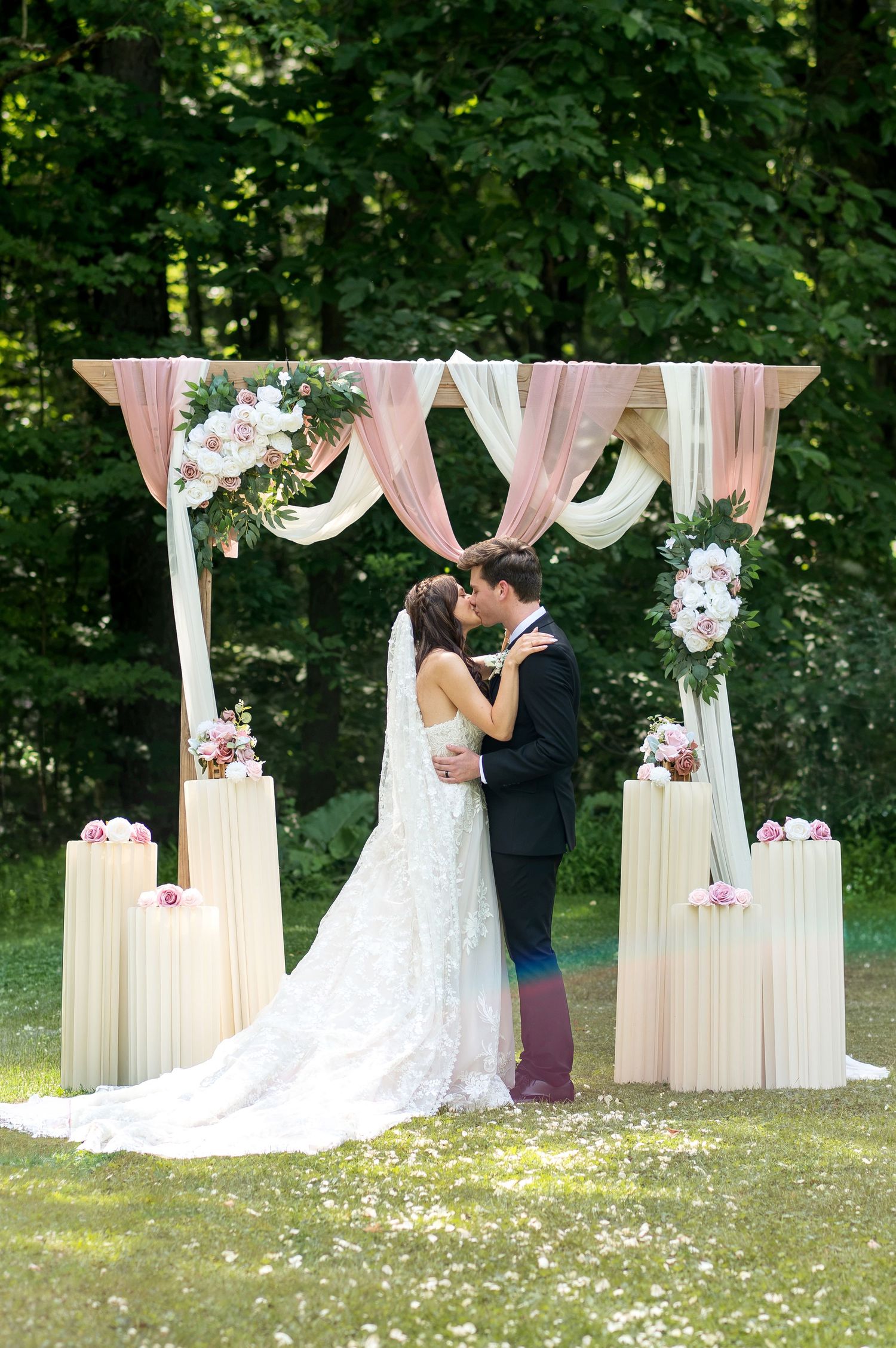 Bride and groom sharing first kiss under elegant floral arch with pink draping and white flowers at outdoor wedding ceremony.