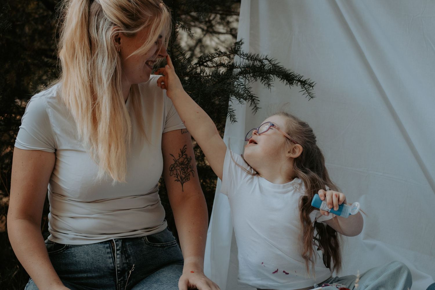 A tattooed mother and her young daughter with glasses share a joyful moment outdoors near a white tent.