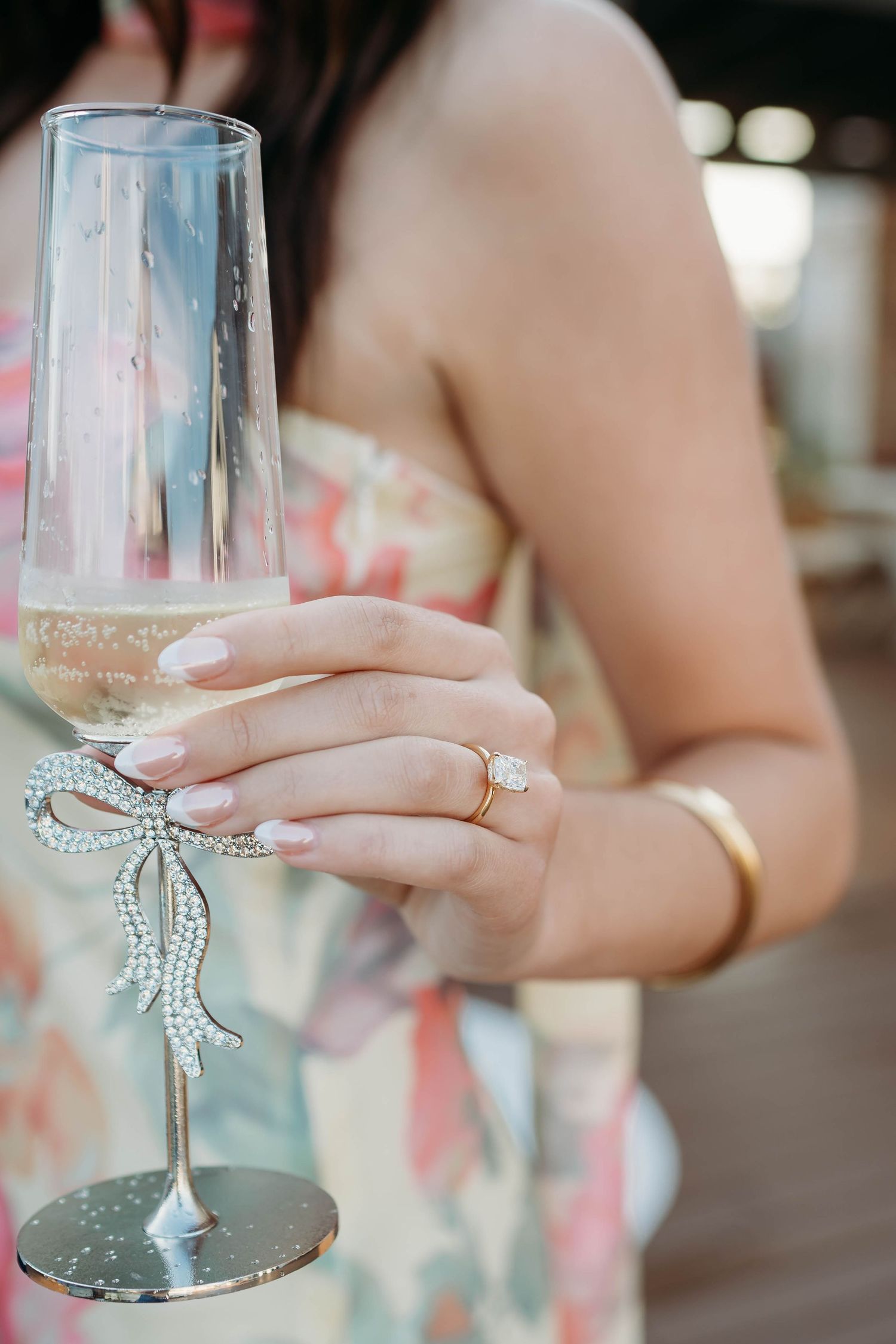 Woman holding a crystal bow champagne flute, wearing a floral dress, diamond ring, and gold bracelet.