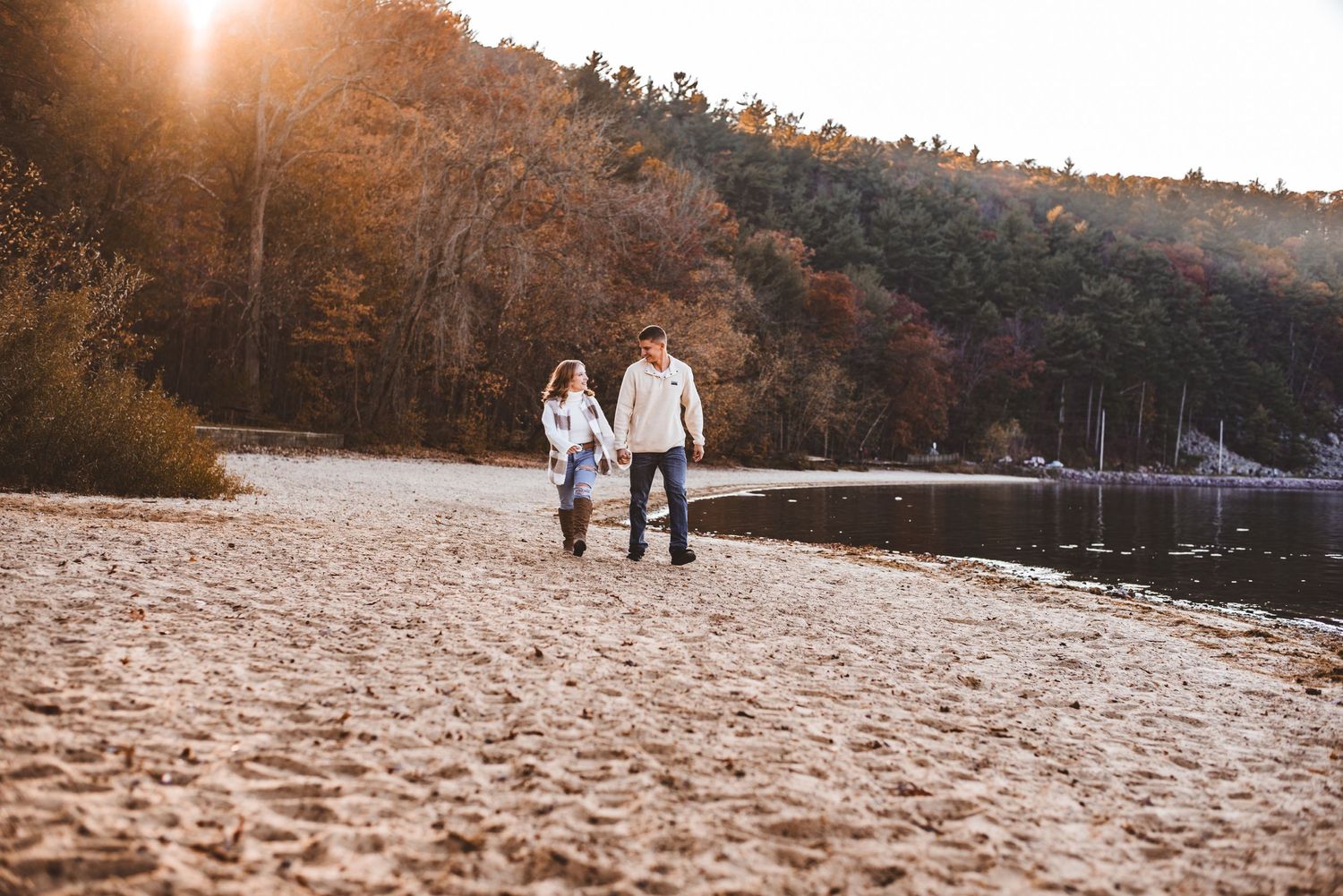 A couple walks along a sandy beach shore at sunset with autumn foliage in the background.