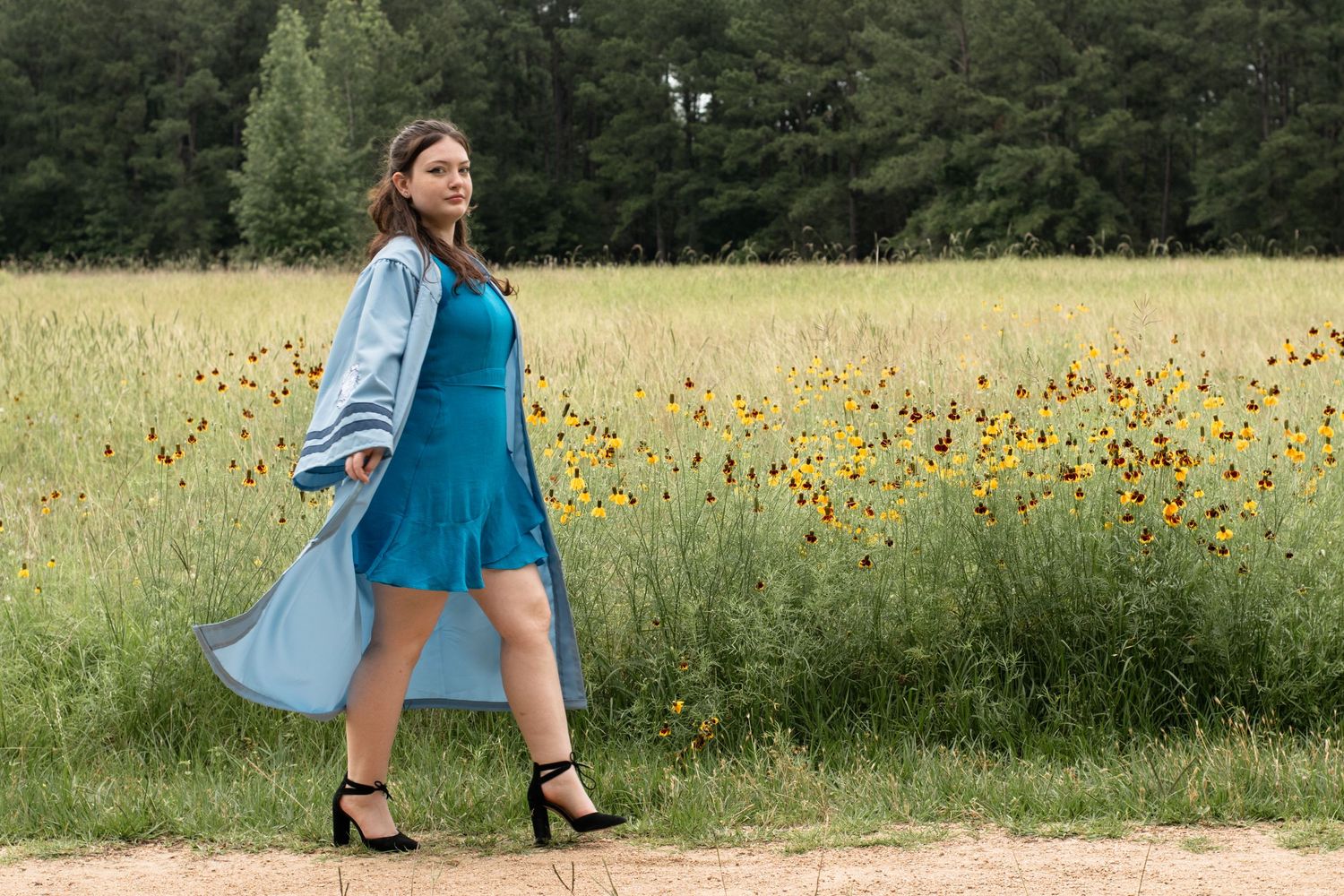 Person walking along path wearing turquoise dress and light blue coat near field of yellow flowers.