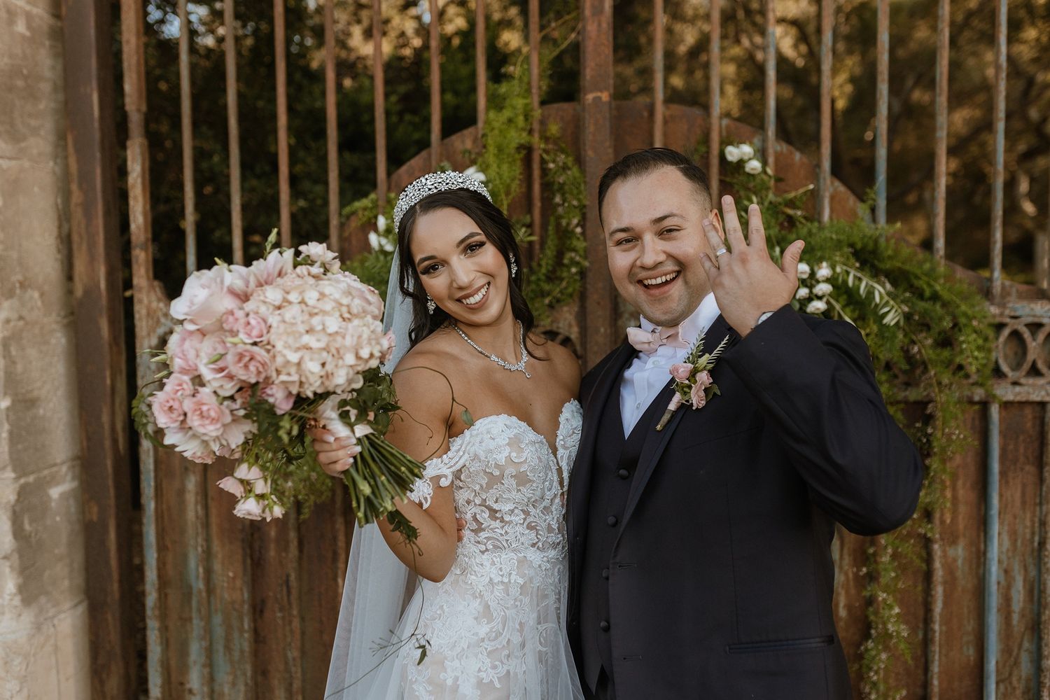A wedding couple poses in front of an ornate iron gate decorated with pink floral arrangements at The Havens Country Club.