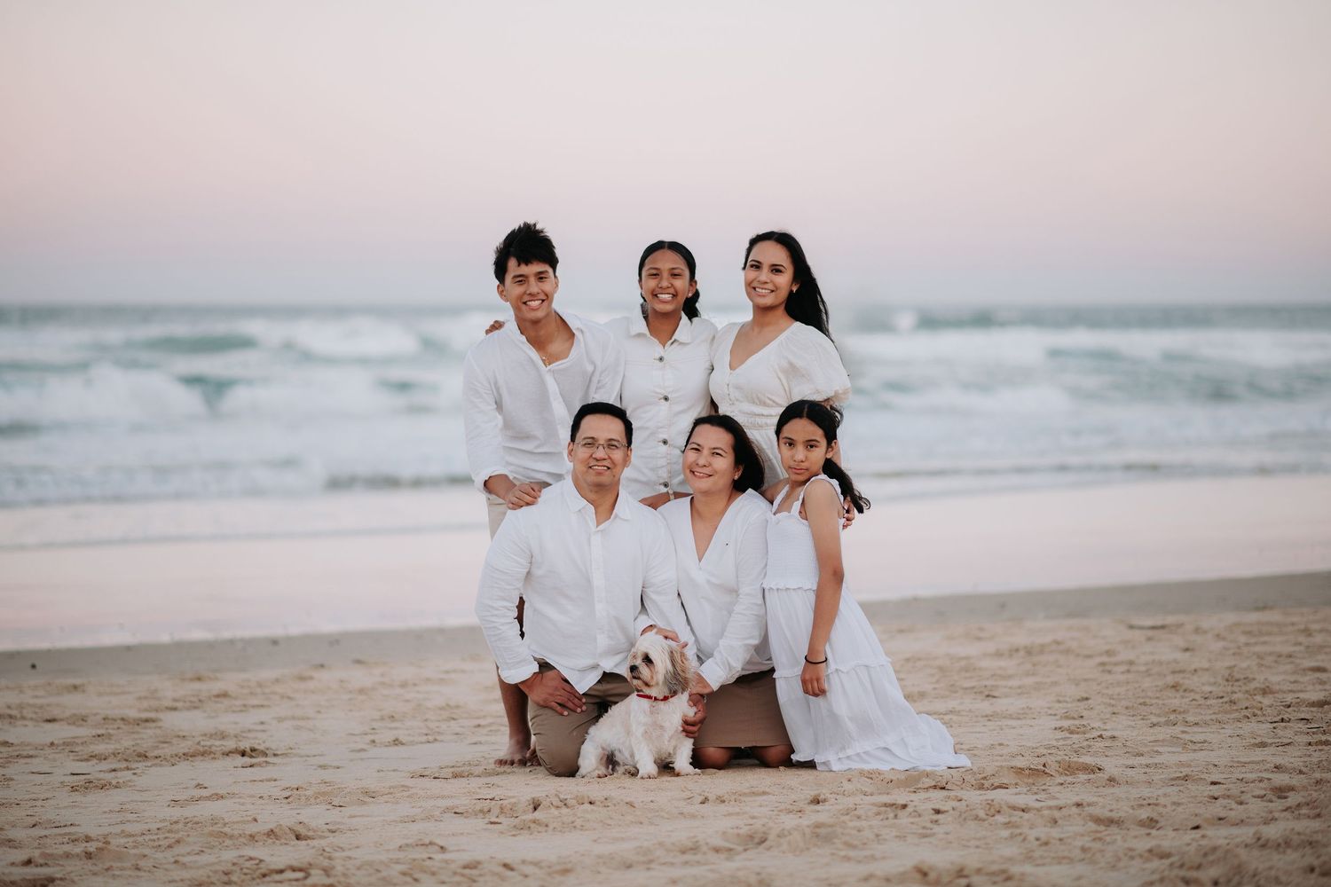 Family in white outfits posing on beach at sunset with their small white dog.