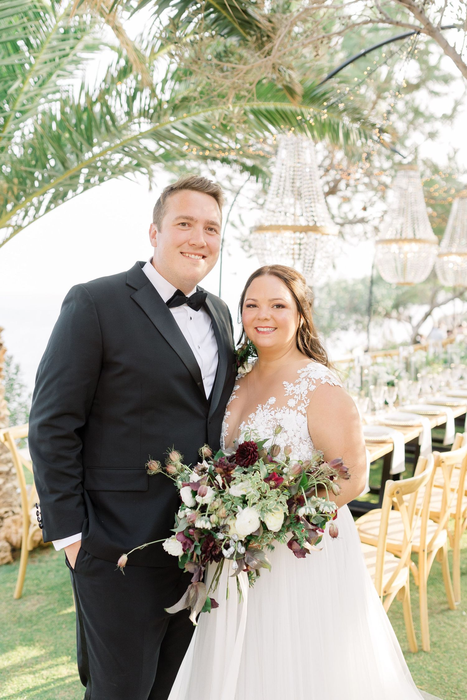 A bride and groom pose together outdoors with palm trees and a floral bouquet at a wedding ceremony.