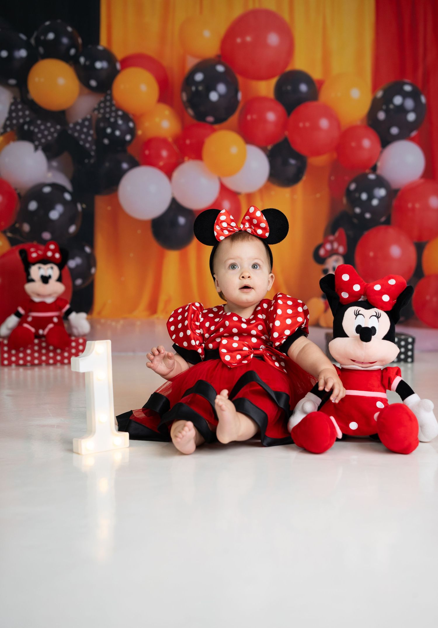 First birthday party setup with red and black polka dot balloons, glowing number 1 candle, and Minnie Mouse plush toys.