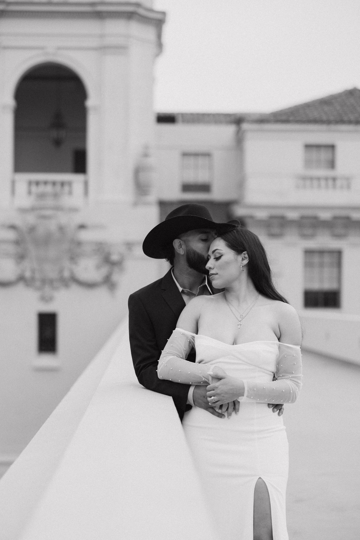 Black and white wedding photo of couple embracing on rooftop, groom in cowboy hat kissing bride.