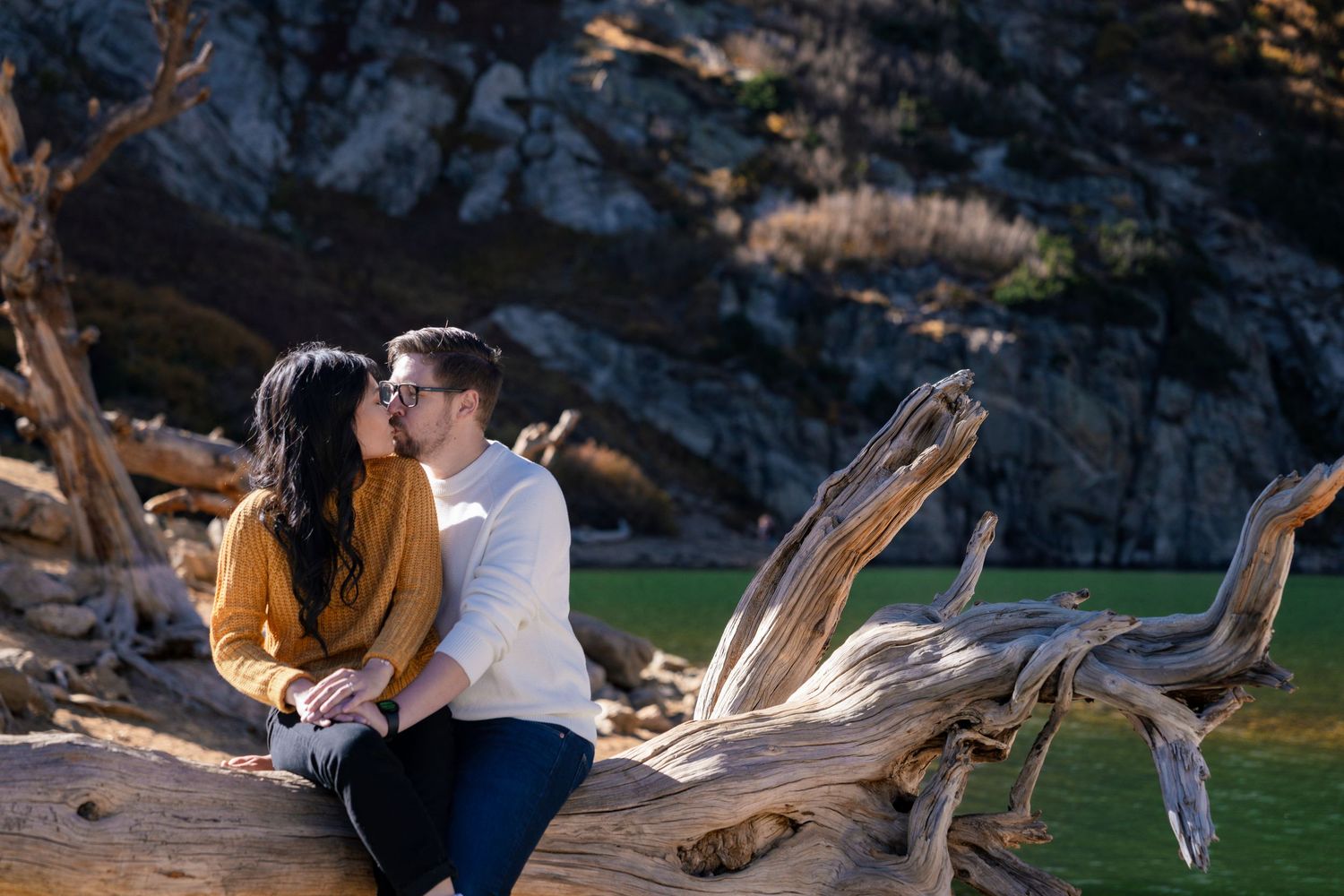 Couple sitting on driftwood at sunset sharing an intimate moment against rocky cliffs in nature.