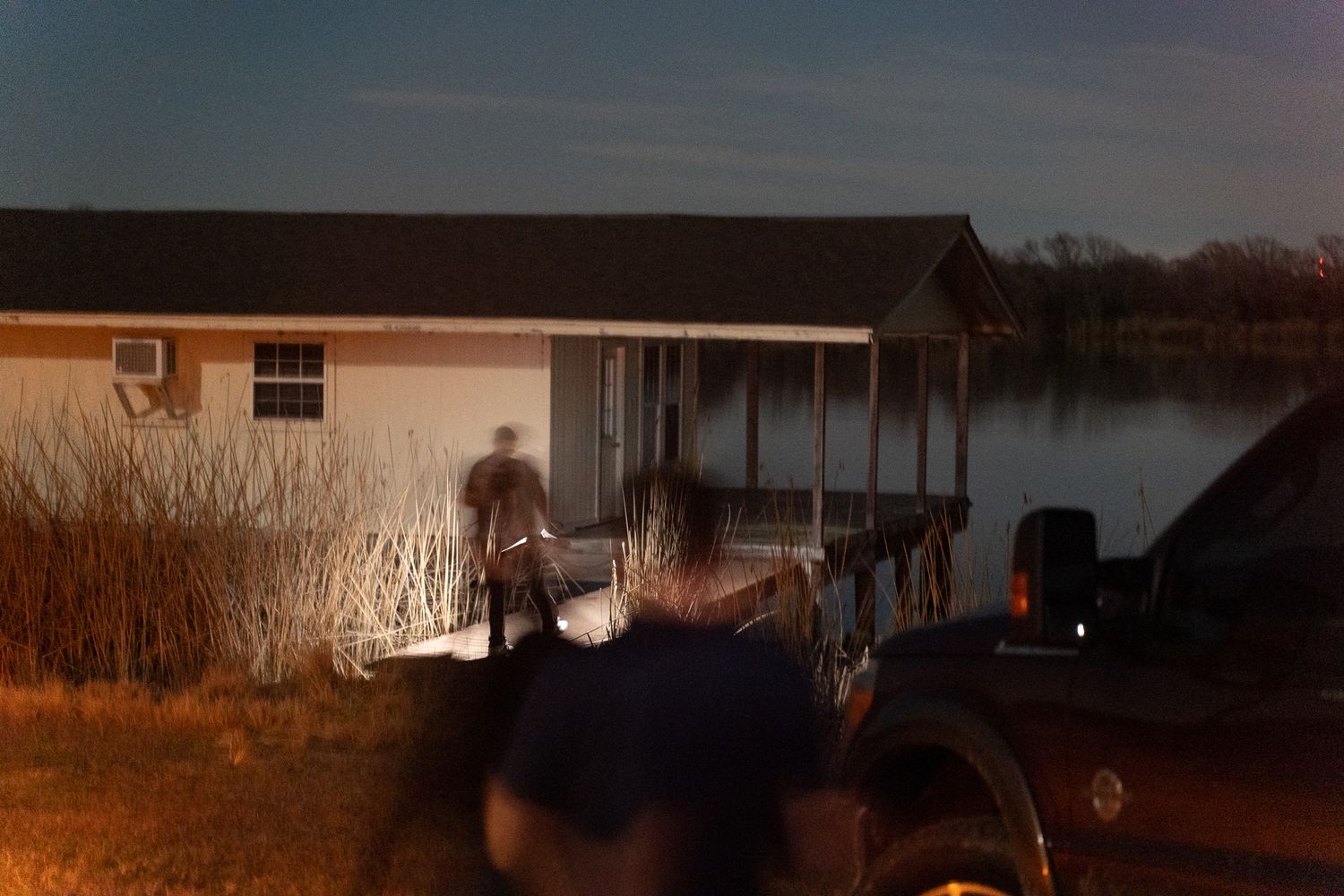 Dark nighttime scene by a lake with silhouettes and dock visible in low light.