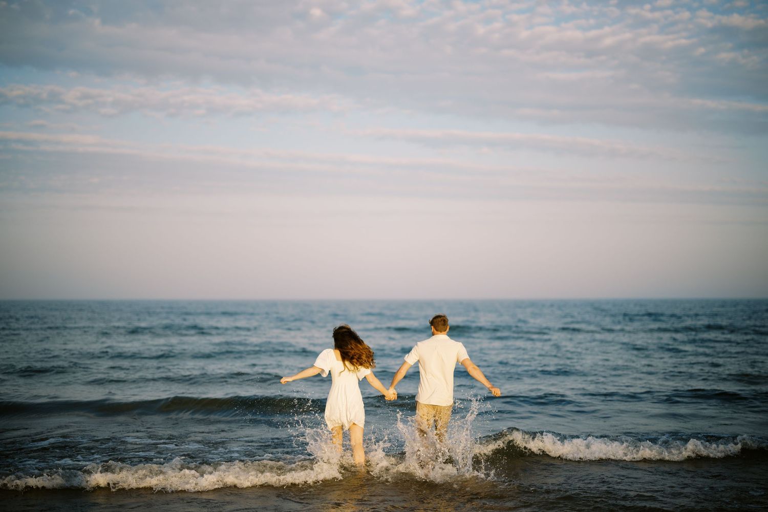 A couple in light colored clothing splashes together in the ocean waves at sunset.