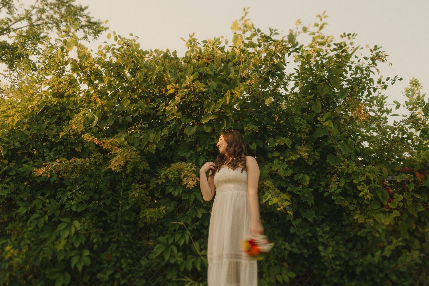 Figure in a flowing white dress stands before lush green foliage at sunset holding vibrant flowers.
