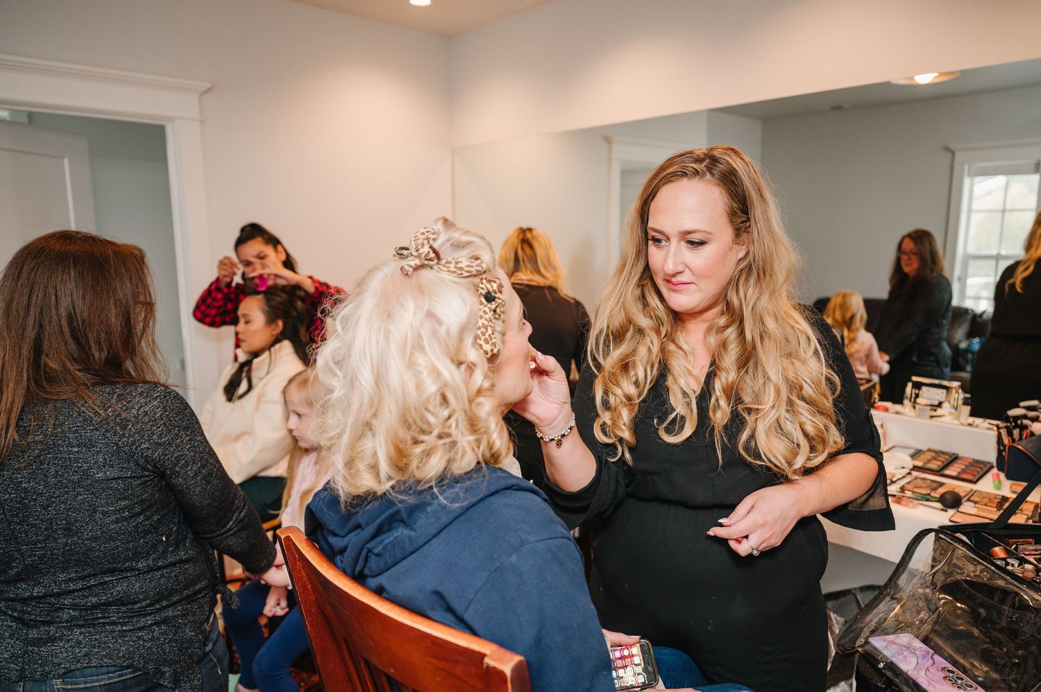 Makeup artist styling client's hair in preparation area with other beauty professionals at work in background.
