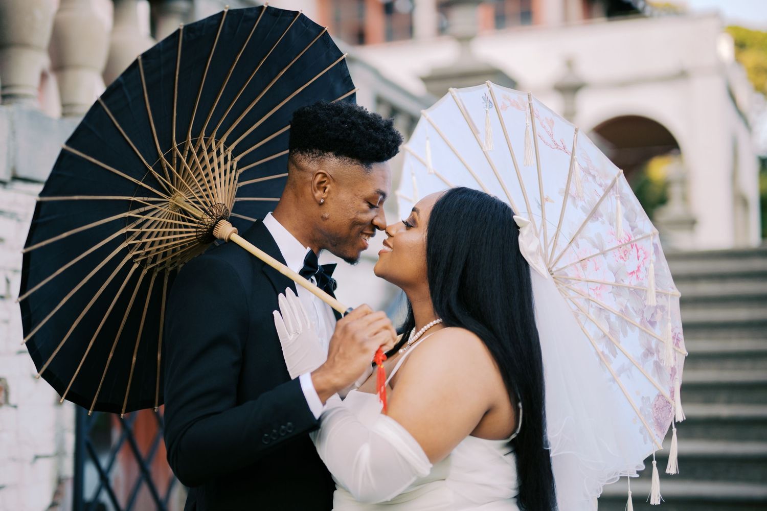 A romantic couple poses with decorative parasols on stone steps, sharing an intimate moment.