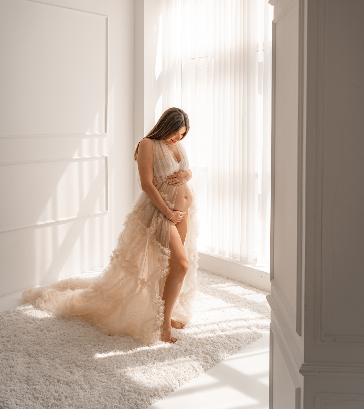Ethereal maternity photo in sheer beige gown with soft sunlight streaming through white curtains in bright room.