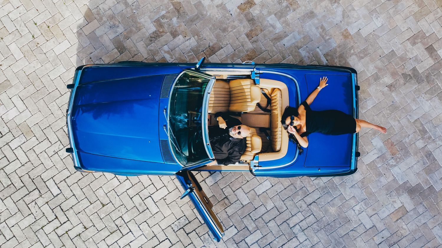Happy couple enjoying a sunny day in a luxury convertible car with tan leather interior and tropical scenery.
