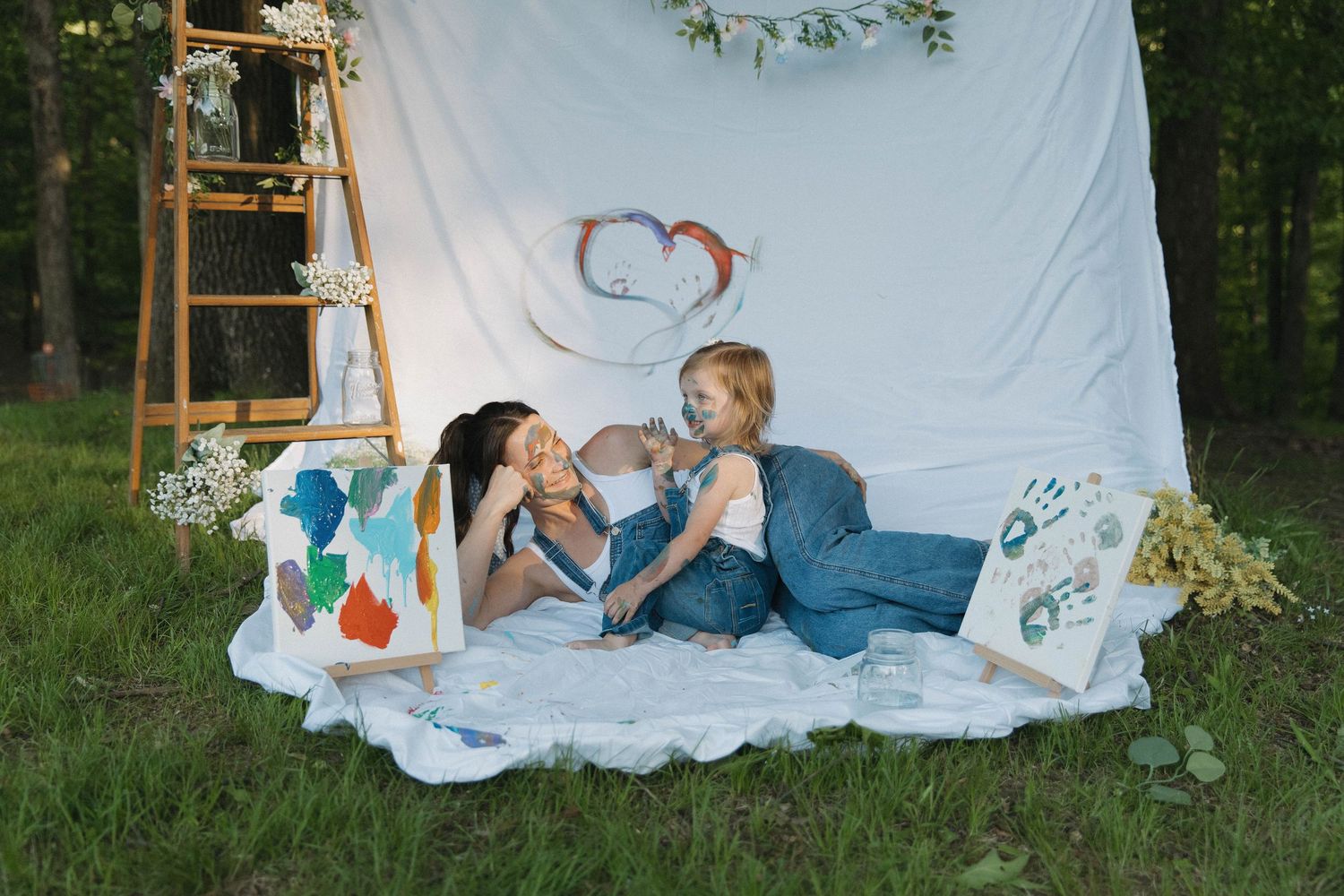Two people lying on a white blanket outdoors with a heart painted backdrop and decorative setup in a woodland setting.