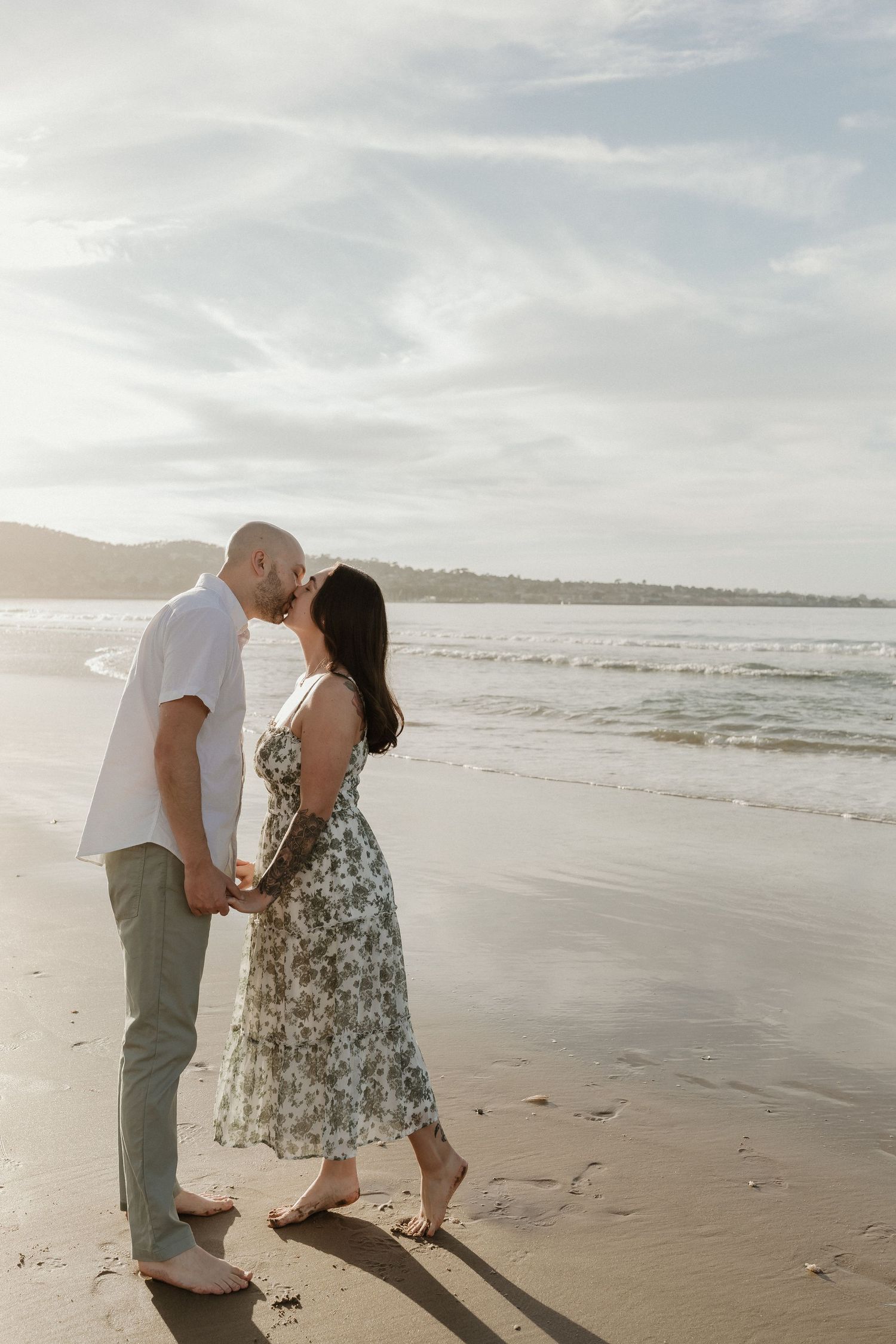 A couple shares an intimate moment while standing on a sandy beach at sunset with ocean waves in the background.