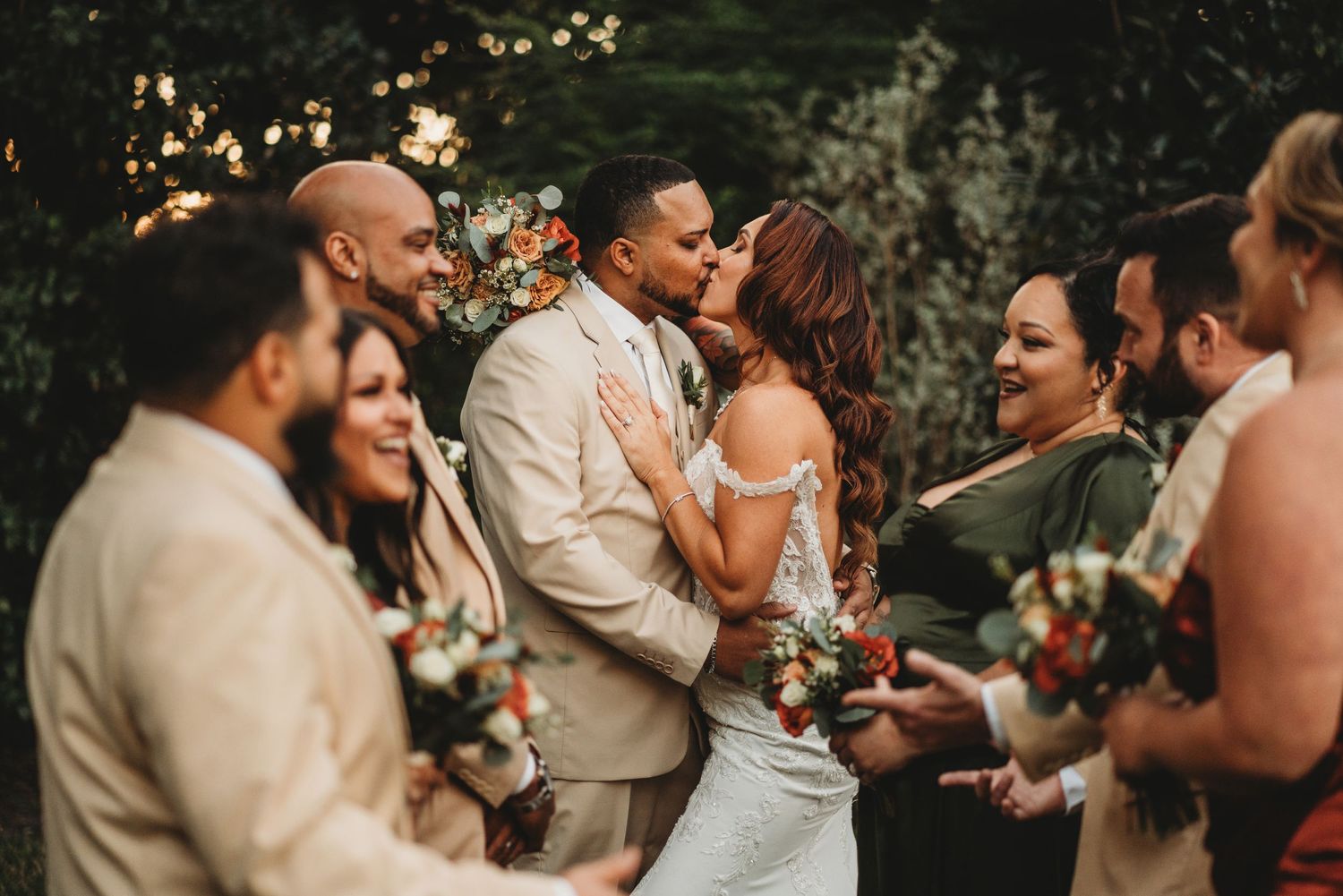 A couple shares a kiss surrounded by their wedding party in an outdoor garden setting.