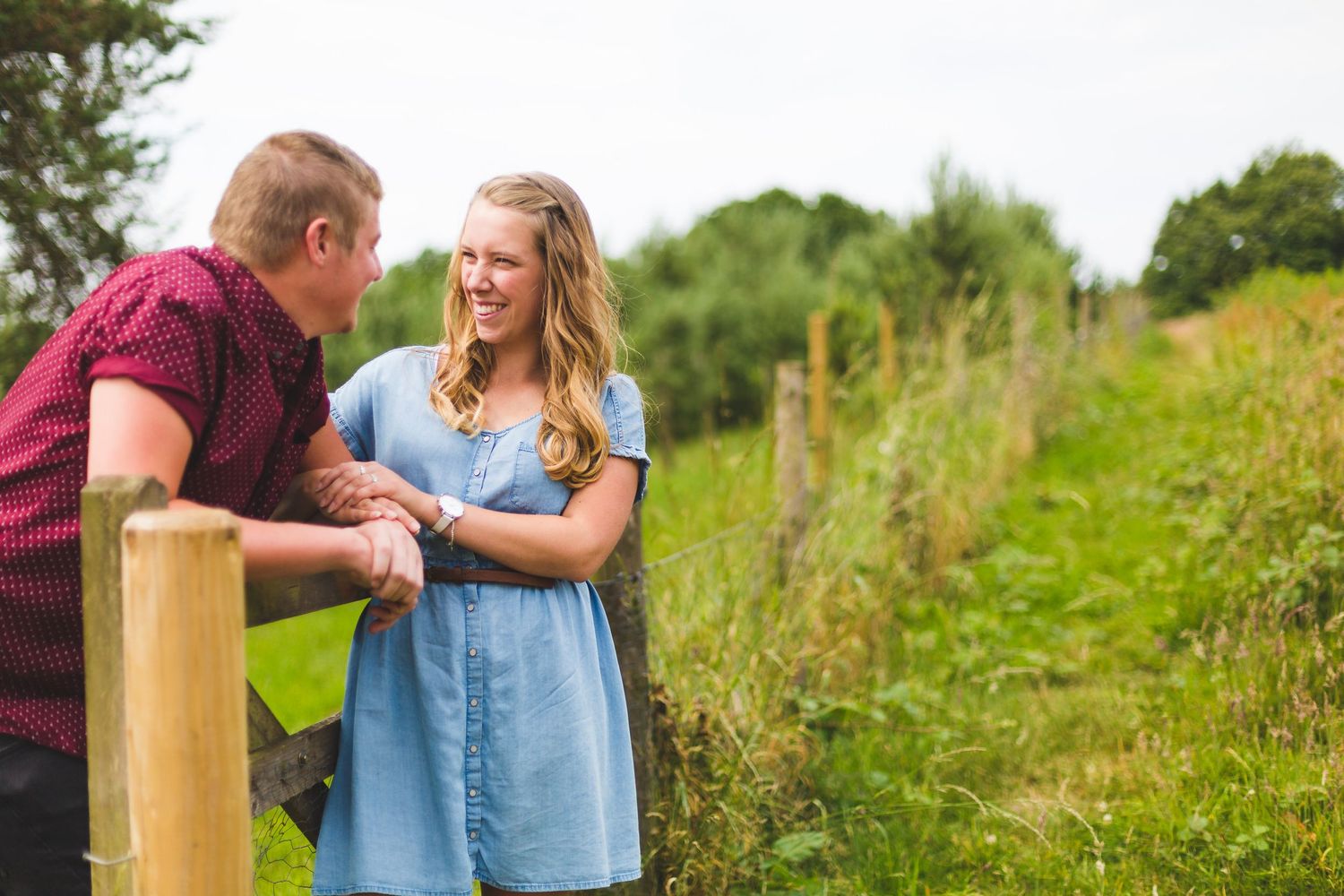 A couple shares a conversation while leaning on a wooden fence post in a rural setting.