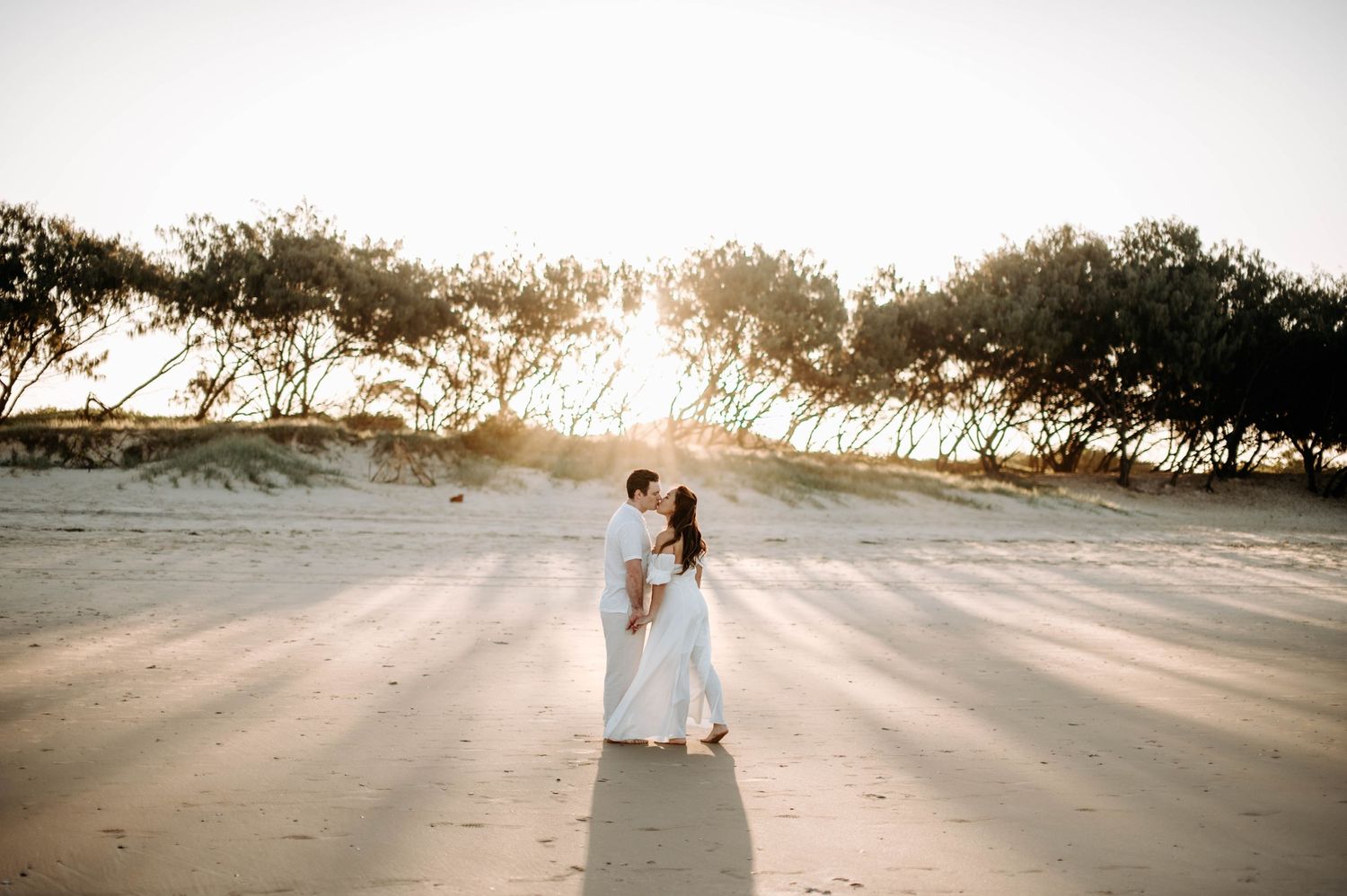 Couple in white outfits poses on beach during golden hour with sunlight streaming through trees.