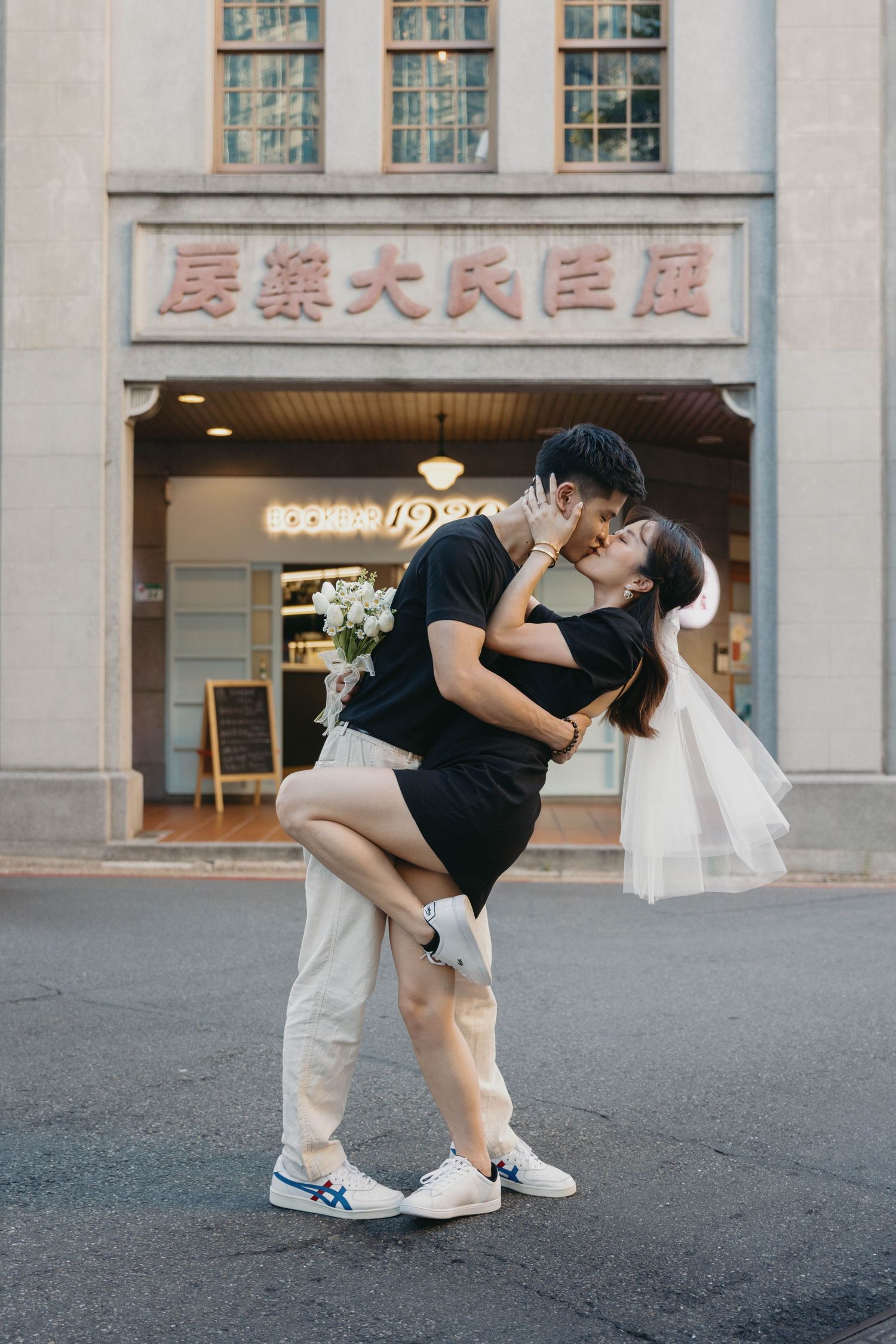 Couple sharing a romantic kiss outdoors in front of a vintage building facade, bride holding flowers.