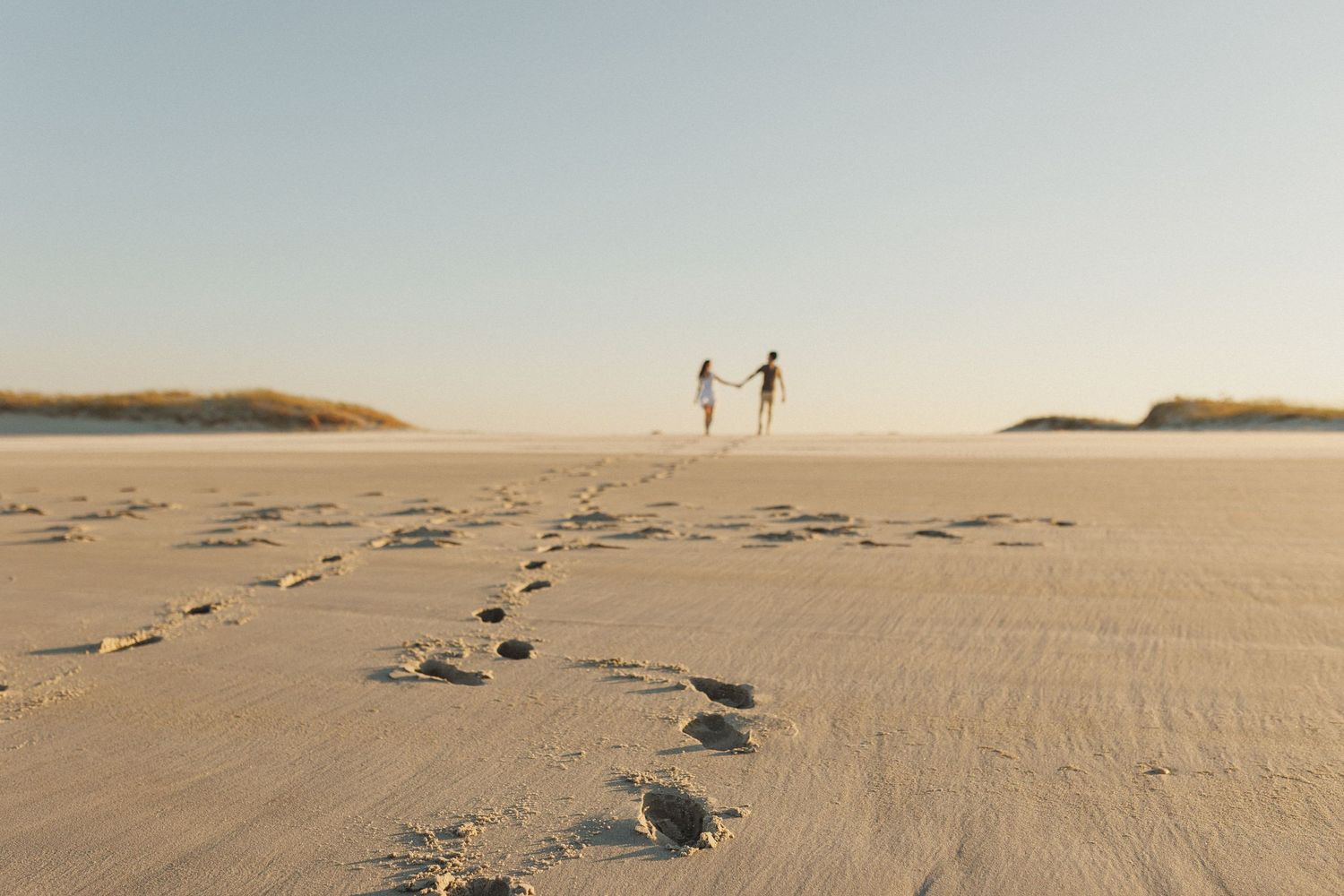 Footprints lead to two distant figures walking along a misty beach at sunset near Lisbon, Portugal
