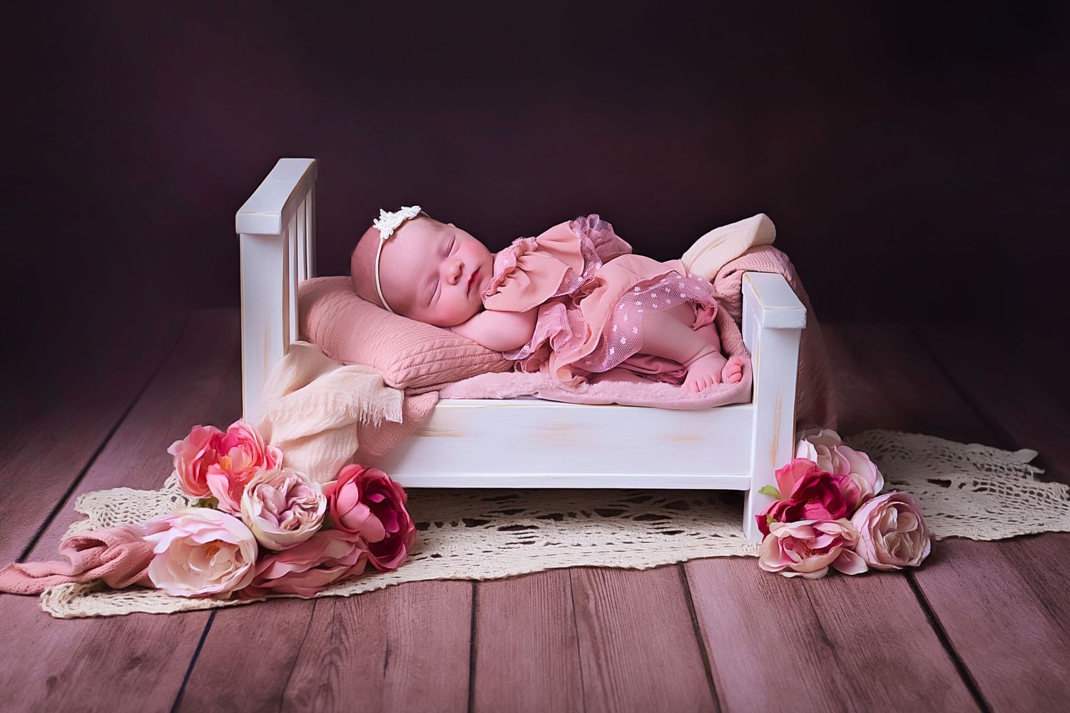 Newborn baby in pink outfit sleeping peacefully on white wooden bed with pink roses scattered on wooden floor.