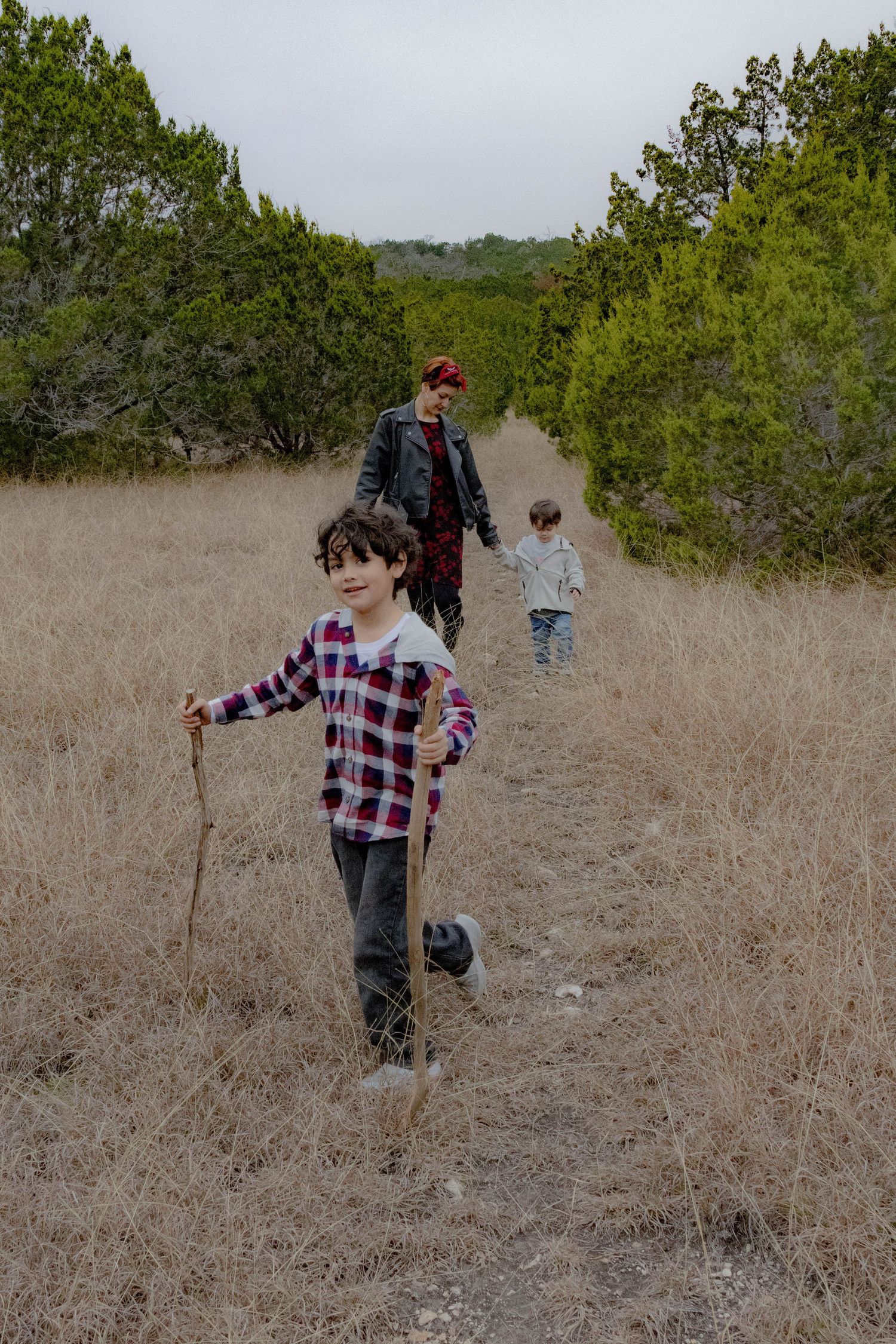 Hikers walk along a dirt trail through dry grass and green shrubs on an overcast day at a nature preserve.