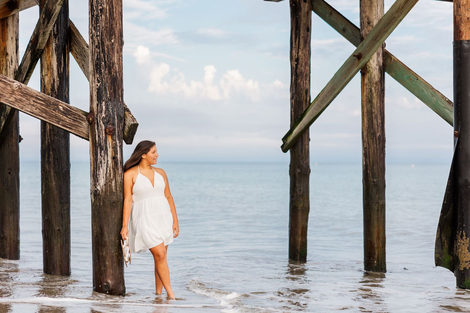 Person in white dress standing between wooden pier posts at oceanfront.